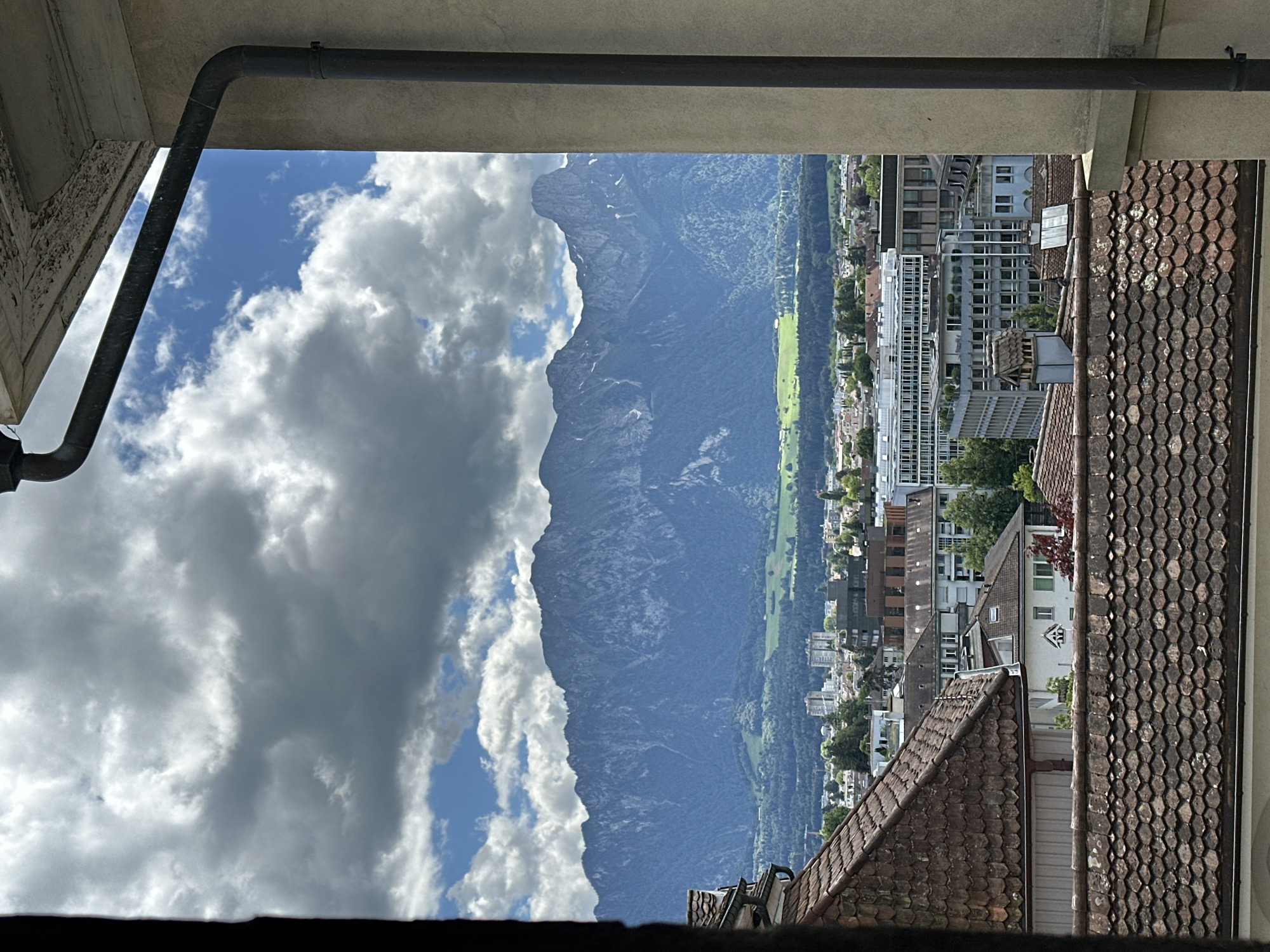 The image presents a view of Thun, Switzerland, from a high vantage point, framed by architectural elements. In the foreground, the weathered, terracotta-tiled roofs of several buildings are visible, some with visible signs of age and moss growth. A vertical, dark-colored drainpipe runs along the right edge of the frame, attached to a cream-colored concrete wall.
Beyond the immediate rooftops, the mid-ground reveals a cityscape characterized by a variety of buildings. These include structures with light-colored facades and numerous windows, some appearing to be modern office buildings or apartment complexes, interspersed with older, more traditional architecture. Green trees are scattered throughout the urban area, suggesting a well-maintained urban landscape.
The background is dominated by a majestic mountain range, its peaks sharp and rocky, some showing patches of snow. The mountains are a deep blue-gray in color, contrasting with the lighter hues of the city and the sky. Lush green fields and forests stretch across the foothills leading up to the mountains, indicating a rural area surrounding the city. The sky is dynamic, with a mix of dark, heavy clouds and patches of bright blue sky, suggesting a changeable weather day. The overall scene conveys a sense of depth, transitioning from the close-up details of the urban architecture to the vastness of the natural landscape. There are no people or active events depicted in the image.