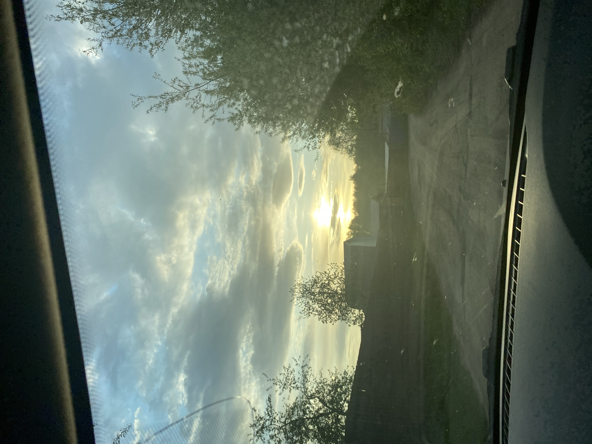 A photograph taken from inside a vehicle, likely a car, looking out through the windshield. The vehicle is on a road in what appears to be a rural or suburban area. The sky is filled with clouds, and the sun is positioned centrally in the frame, causing lens flare. Trees are visible on both sides of the road. Buildings, possibly houses, are visible in the background. The scene appears to be taken at sunset or sunrise. Without further information, the specific location in Chaikovsky, Russia, cannot be determined.