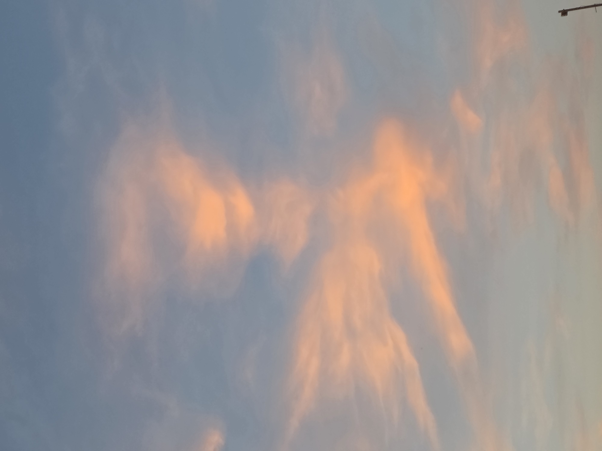 The image shows a light blue sky with orange clouds. The clouds are concentrated in the center and right of the image, appearing wispy and layered. A dark object is visible in the upper right corner. The image is rotated, and no information suggests its location is Hurghada, Egypt. There are no people visible.