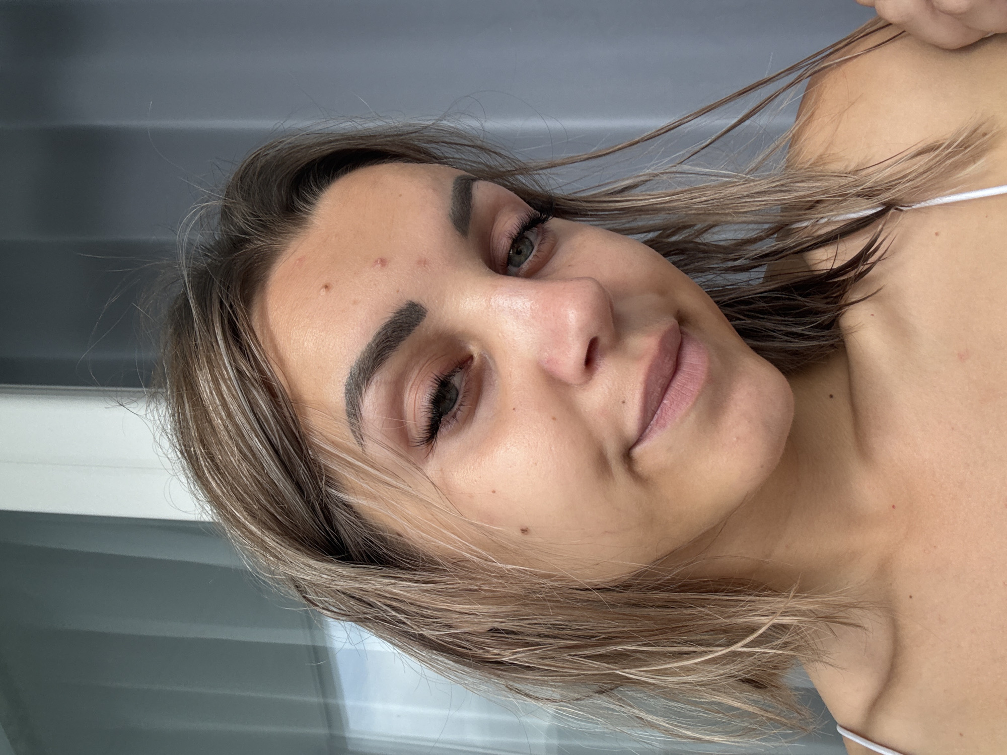 A young woman with blonde, shoulder-length hair that is wet and slightly messy, is looking directly at the camera. She has light skin with some blemishes and a small mole on her cheek. Her eyebrows are dark and well-defined, and she is wearing subtle eye makeup. Her lips are slightly parted, and she has a faint smile. She is wearing a white camisole. The background appears to be an indoor setting with a window and a grey curtain. The lighting suggests it is daytime. This image was taken in the city of Kholmsk, Russia.