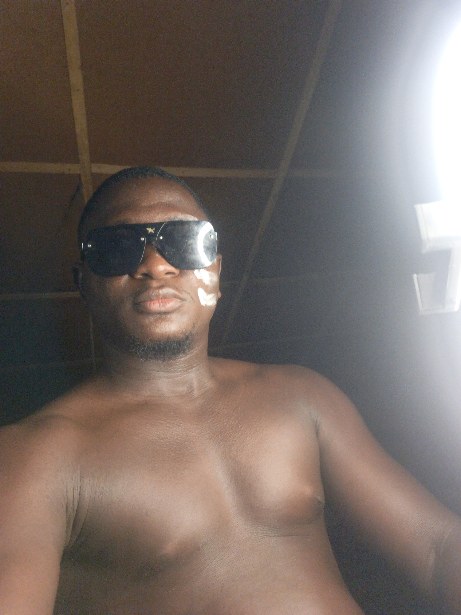 A shirtless, dark-skinned man with a short beard and sunglasses poses for a selfie. He has some white markings on his left cheek. The setting appears to be indoors with a ceiling made of brown boards with light-colored beams. There is a bright light source to the right. The location is assumed to be Akwanga, Nigeria.