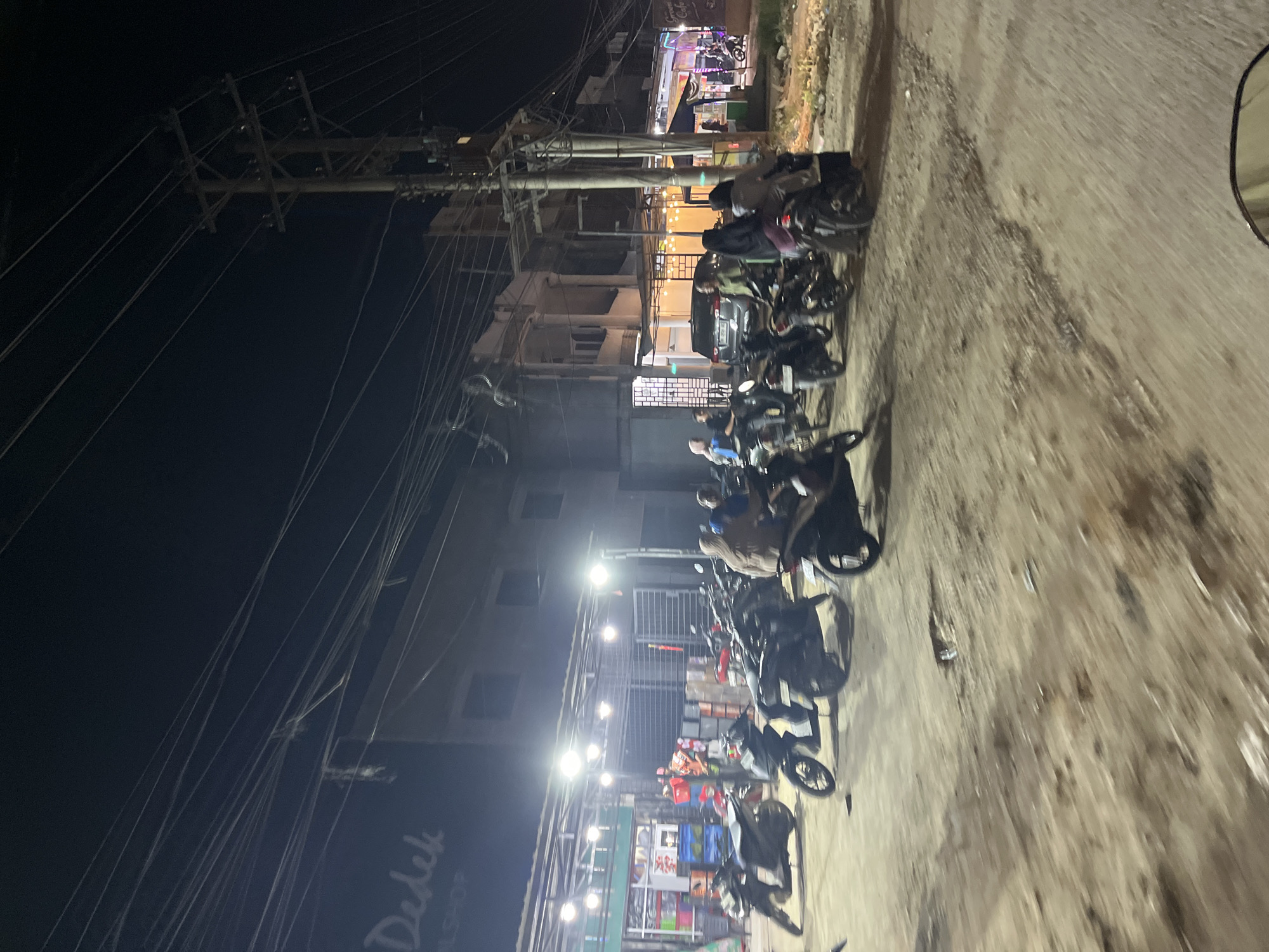 The image depicts a nighttime scene in what appears to be a commercial area or street in Bundar, Indonesia. The primary subjects are a line of parked motorcycles, with some people seated on or standing near them. In the background, there are buildings with some illuminated windows and a sign that reads "Dedek BSNL SHOP". The setting is characterized by the presence of numerous utility poles with tangled wires, suggesting an urban or semi-urban environment. The ground is a dirt or gravel surface, showing signs of wear. The lighting is artificial, coming from streetlights and building lights, indicating it is nighttime. There is no clear indication of weather, but the ground does not appear wet. The overall mood is one of everyday activity, likely people gathering or taking a break at a shop or establishment. The image's orientation is portrait, but the content is captured in landscape format, suggesting the photo was taken from a moving vehicle, possibly a motorcycle, looking towards the side.