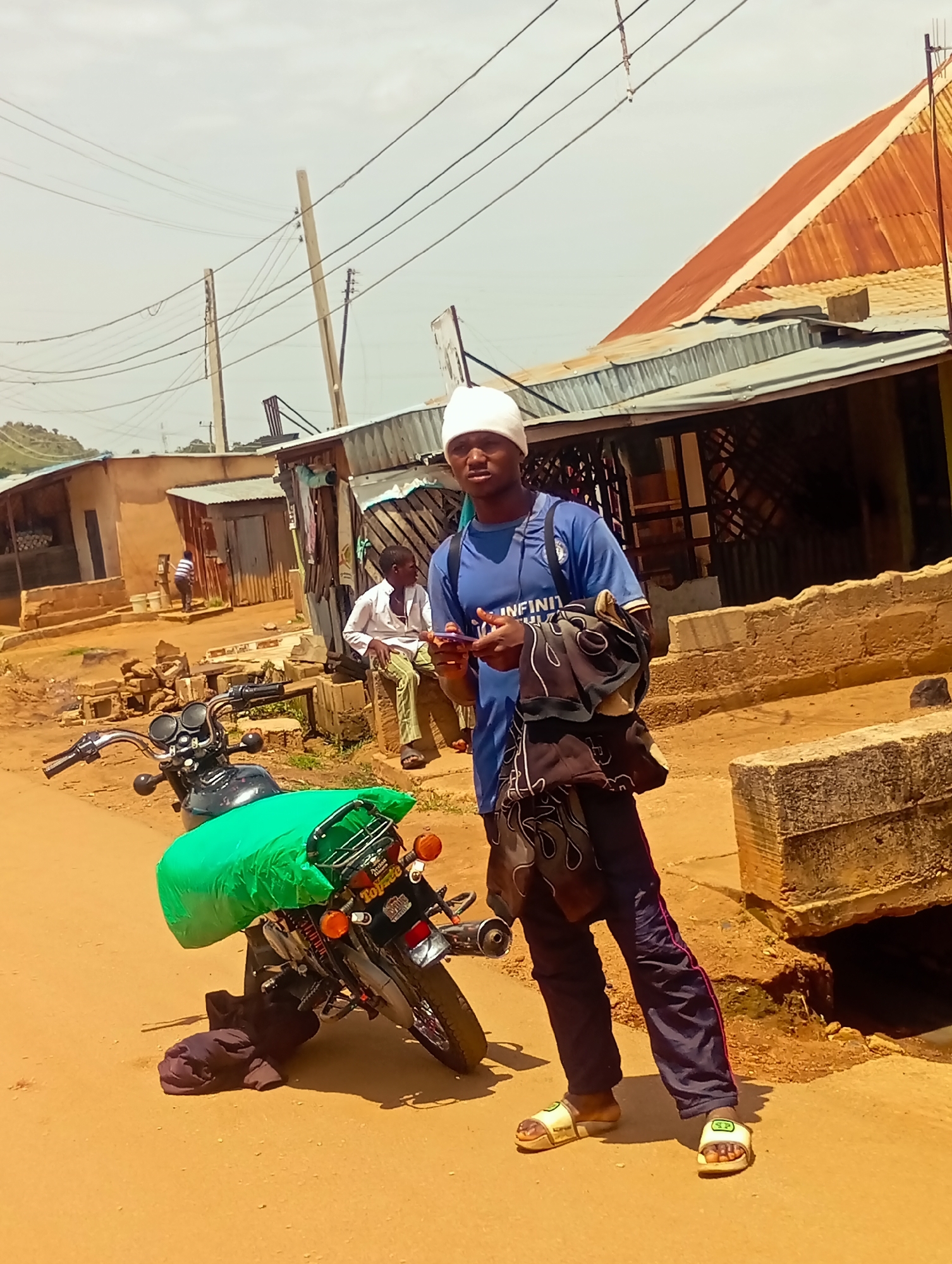 In Du, Nigeria, a young man wearing a white kufi and a blue t-shirt stands next to a black motorcycle. The motorcycle is loaded with a large green tarp covering its rear. He is holding a mobile phone in his right hand. He is also carrying a dark bag draped over his left shoulder, with a patterned garment. The man is wearing dark pants with a pink stripe and white sandals. In the background, a boy in a white shirt and green pants is seated on a concrete block. Several corrugated metal structures and a brick building are visible. Overhead, numerous power lines crisscross the sky, supported by poles. The ground is a dusty, orange dirt road.