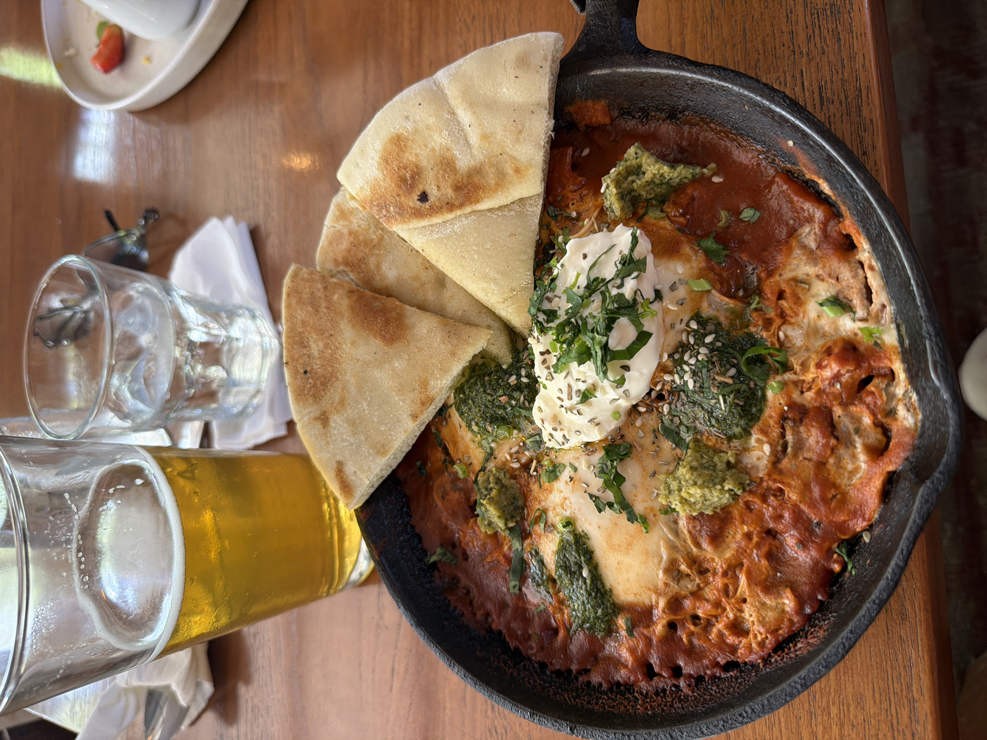 On a wooden surface, a cast iron skillet holds food, accompanied by pita bread pieces. A glass of amber liquid and a clear glass sit nearby. The food in the skillet appears to be eggs in tomato sauce, topped with a white dollop and sprinkled herbs and seeds. A small white plate with a red fruit slice is also present. The city and location are unknown.