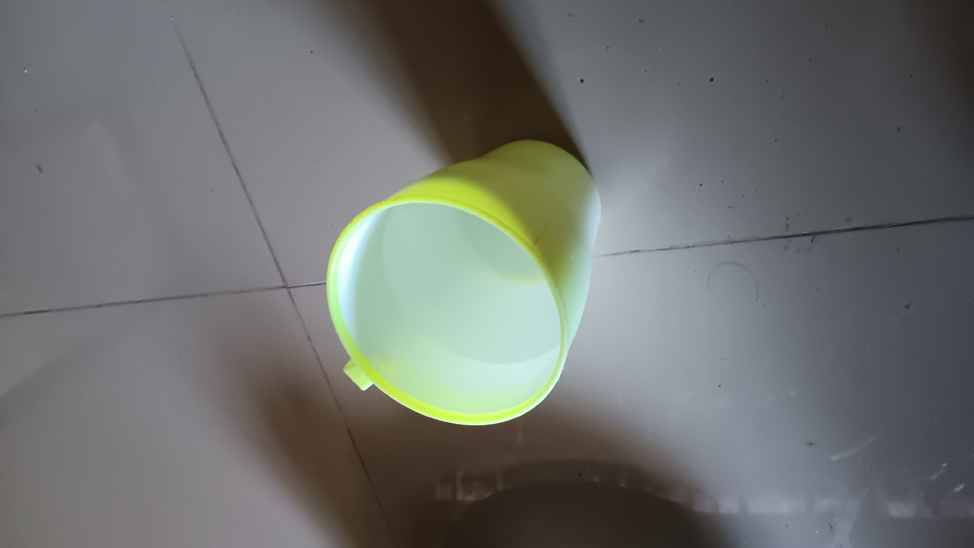 A pale yellow plastic cup filled with water sits on a tiled floor. The cup is positioned at an angle, with its opening facing upwards and slightly to the right. The water level appears to be about halfway up the cup. The floor is made of light-colored square tiles, with dark grout lines visible between them. The lighting suggests an indoor environment, possibly a home or public space, with a shadow cast by the cup and an overhead light source. There is no indication of the specific location within Sonobekel, Indonesia, beyond the general description of the environment. No people are present in the image.