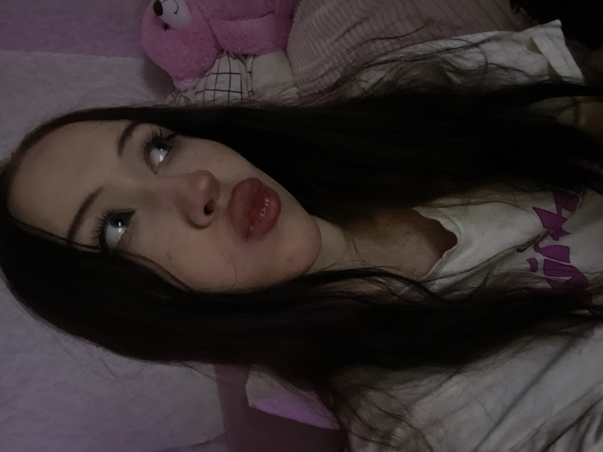 The image is a selfie of a young woman with long, dark hair and light-colored eyes. She is wearing a white t-shirt with a purple design on it. Her lips are puckered, and she is looking off to the side. Behind her, a pink teddy bear is visible, along with a pink blanket and a checkered pillow. The lighting in the image is dim and appears to be from a purple light source, possibly a lamp. The background suggests an indoor setting, likely a bedroom. There is no discernible information within the image that would place it in Votkinsk, Russia.