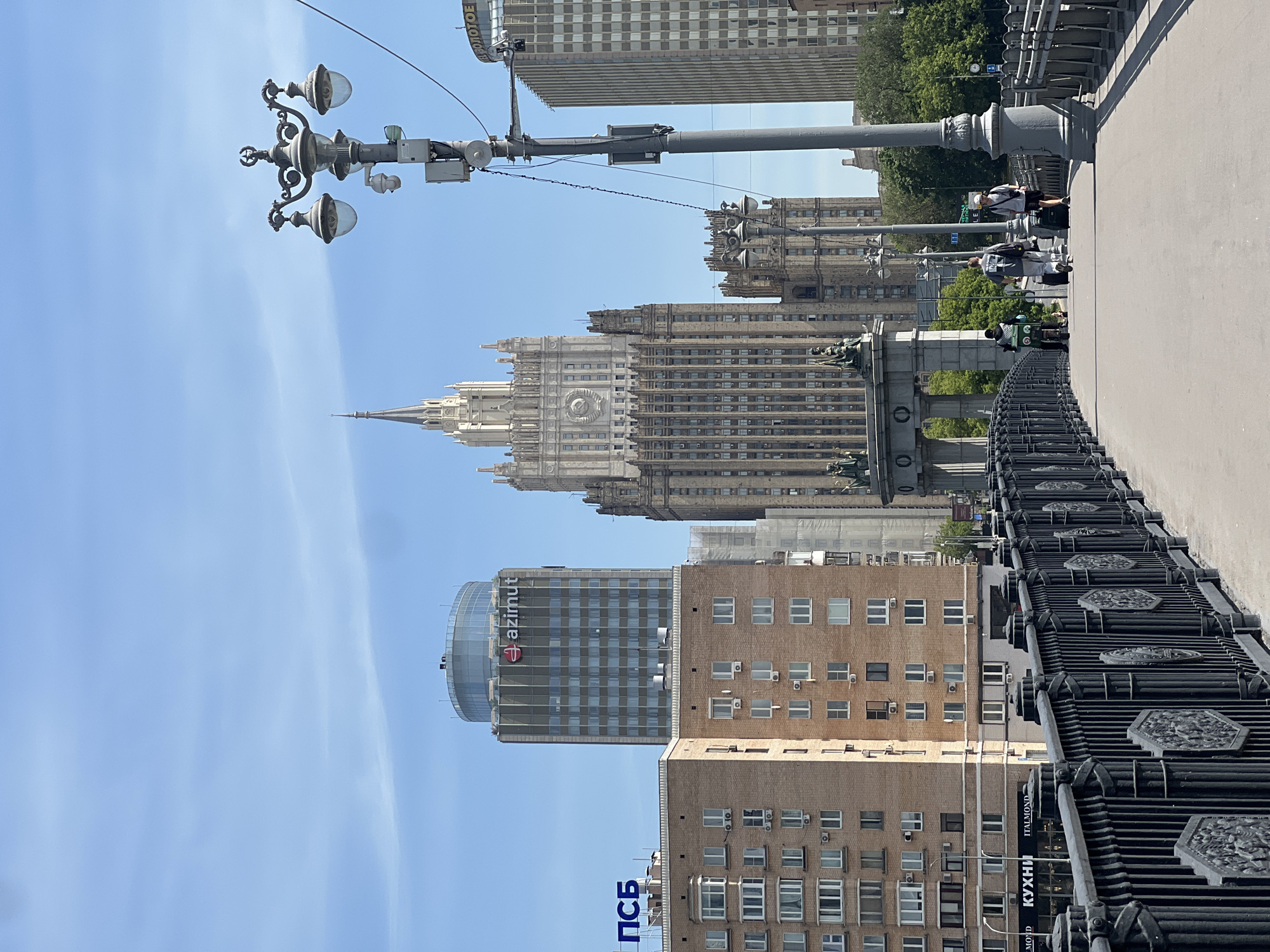 The image depicts a daytime scene on the Borodinsky Bridge in Moscow, Russia, offering a view towards the city's architectural landmarks under a clear blue sky with sparse wispy clouds.
In the foreground, a wide, light-colored pedestrian pathway of the bridge extends away from the viewer. To the left, a dark, ornate metal railing with decorative hexagonal panels runs along the path. To the right, a tall, dark gray lamppost with an elaborate three-pronged light fixture is visible, featuring a CCTV camera and several electronic boxes attached to its pole.
Three individuals are walking away from the viewer on the pathway. The leftmost person is riding a bicycle and carrying a large, green delivery bag on their back. The middle person, a male, is dressed in light clothing and pulling a black wheeled suitcase. The rightmost person, also a male, wears a white cap, dark top, and shorts, and is likewise pulling a black wheeled suitcase. A smaller, more distant figure is faintly visible further down the path.
In the midground and background, several prominent buildings define the Moscow cityscape. The most dominant structure is the Ministry of Foreign Affairs building, a large Stalinist Empire-style skyscraper characterized by its light-colored facade, a central spire, and a large clock face adorned with a Soviet emblem (hammer and sickle within a star wreath) below it. To the left of this building stands a modern, taller building with a cylindrical glass top, featuring the "azimut" (Azimut Hotels) logo in red letters near its roof. Below the Azimut building, a brick-colored building with multiple windows and visible air conditioning units bears a blue "ПСБ" (PSB) sign on its roof. To the right of the Ministry of Foreign Affairs building, another tall, light-colored building with a grid-patterned facade shows a partial sign "ВОЛОТОЕ" near its top. Green trees are visible at the base of the buildings and along the right side of the scene.