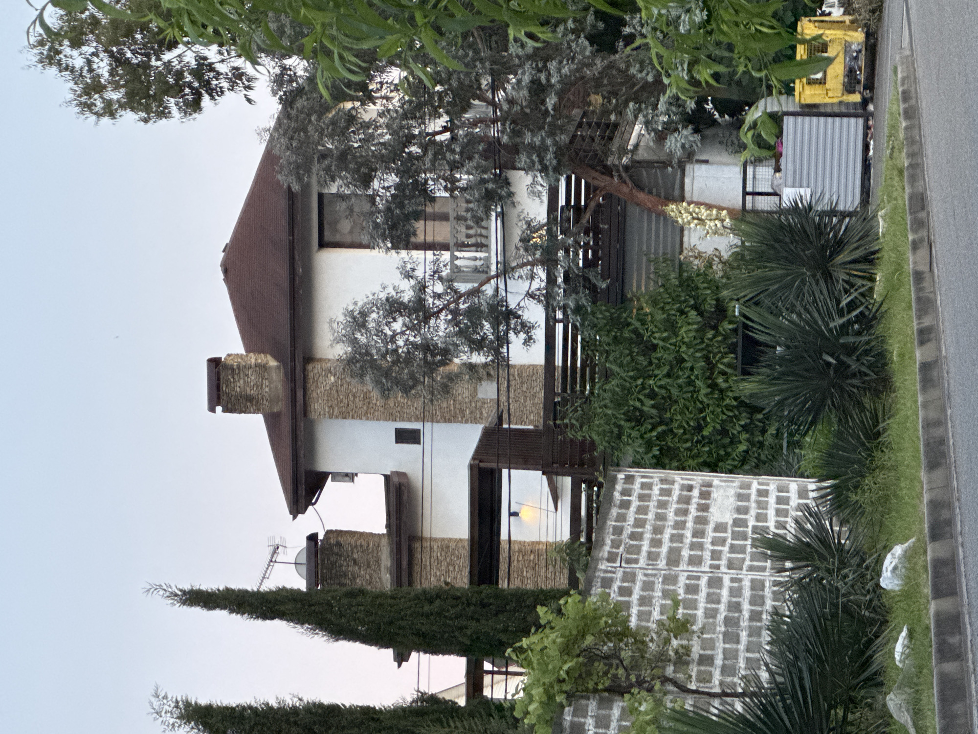 The image displays a multi-story residential building situated in a lush, subtropical environment characteristic of Sochi, Russia. The building features white stucco walls contrasting with substantial natural stone cladding on its chimneys and accent sections. The roof is dark reddish-brown, possibly tiled. On the upper level, a dark-framed window is visible, and below it, a small balcony with a white stone balustrade. Lower levels feature dark wooden railings or pergolas, with one yellow light emanating from underneath a covered section. A satellite dish and a television antenna are mounted on a chimney on the left side of the roofline.
The foreground consists of an asphalt road with a grey and black striped curb. Immediately behind the curb is a grassy verge interspersed with several light-colored rocks. A grey concrete block retaining wall, featuring a repeated square pattern, stands on the left. In front of the wall and throughout the lower-left and central areas are various green plants, including tall, slender cypress-like trees, and several spiky, palm-like plants. To the right, dense green foliage, including trees with lighter-colored leaves (possibly eucalyptus), partially obscure the house. A large plant with white floral spikes is visible on the right. On the far right, a corrugated metal enclosure partially screens a yellow industrial waste bin or container.
