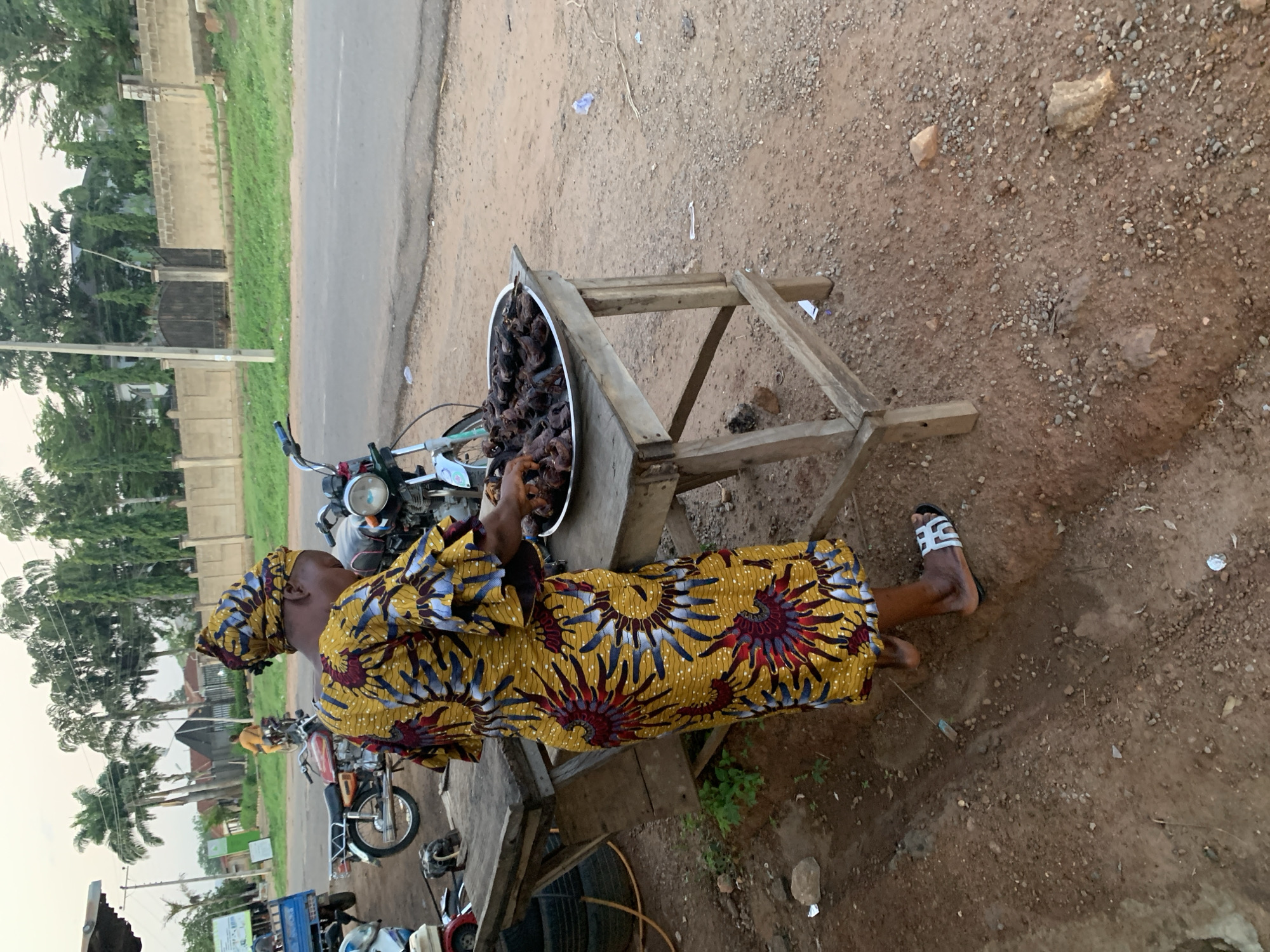 A woman in a yellow dress stands next to a wooden table holding a silver bowl filled with dark-colored meat. The woman is on the side of the road in Gbeleko, Nigeria. Several motorcycles are parked nearby. The ground is dirt and gravel.
