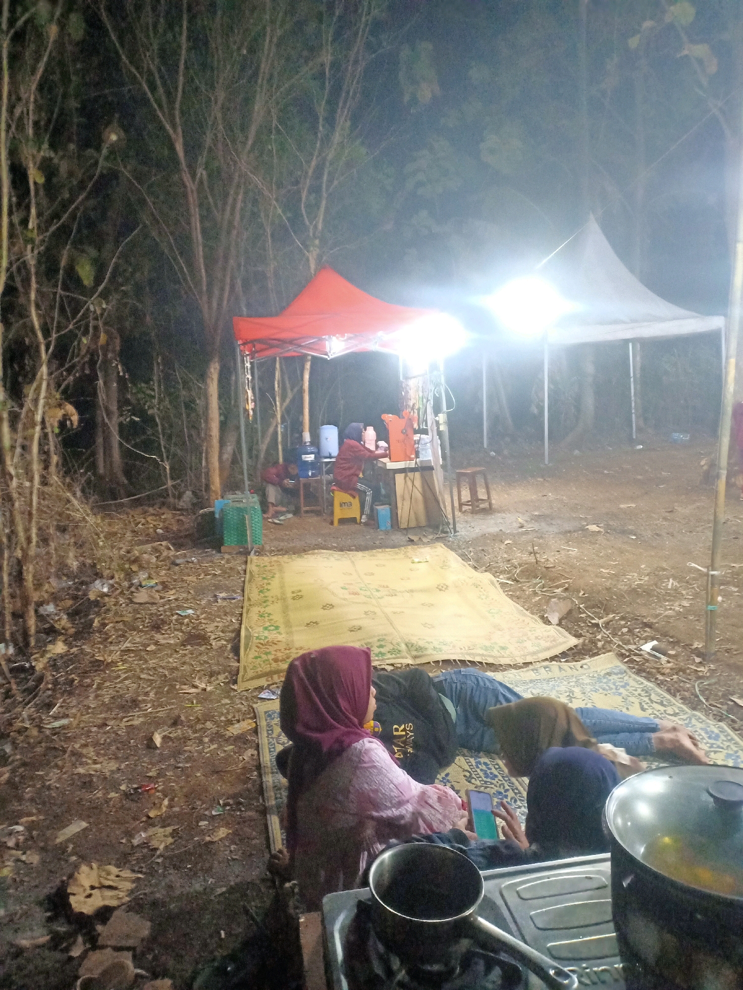 The image depicts an outdoor gathering at night, likely in a forested or rural area in Jatisrono, Indonesia. Two tents are visible, one red and one white, providing shelter and illumination from bright lights. Several individuals are present, some seated and others lying down on mats laid out on the ground. One person in the foreground is wearing a pink headscarf and holding a mobile phone. Another individual is lying down wearing a black t-shirt and blue jeans. The ground is covered in dirt, dry leaves, and scattered debris. Cooking equipment, including a stove and pots, is also visible, suggesting that food may be prepared or consumed at this location. The overall atmosphere appears to be informal and relaxed, possibly a camping or community event.