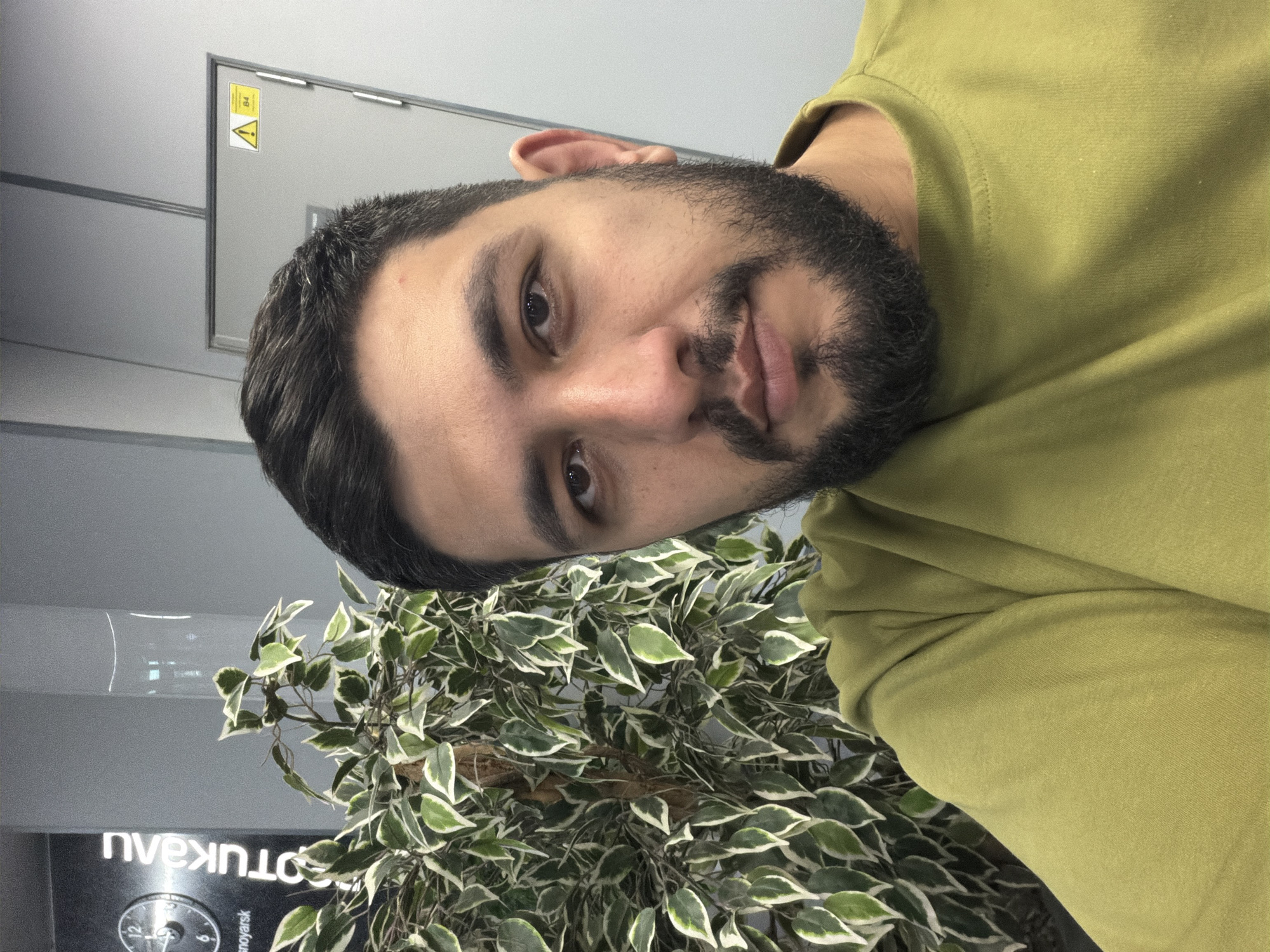 A selfie taken inside a business in Krasnoyarsk, Russia. The person is wearing a green t-shirt. To the left is a variegated ficus plant. Behind the plant is a wall with the business' name on it. A clock and the location "Krasnoyarsk" can be made out. Above that is a utility door with a yellow warning label.