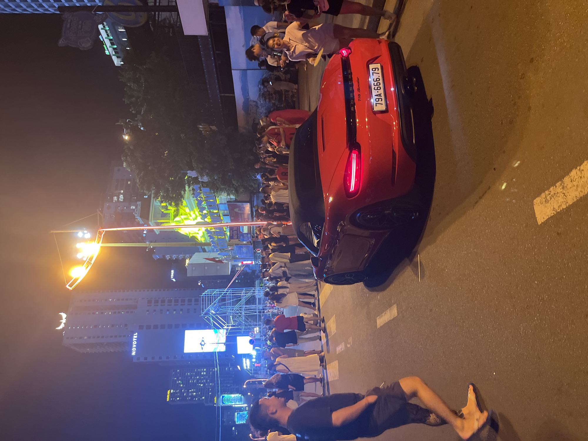 In Nha Trang, Vietnam, a red Porsche Boxster convertible is parked on a street at night, its brake lights illuminated. A man wearing a black t-shirt and dark shorts stands on the left side of the street, looking to his right. Behind the car, a crowd of people, dressed in casual summer attire, are gathered on the sidewalk and street, suggesting a festive atmosphere. In the background, tall buildings, including the Novotel hotel, are visible, adorned with lights. The street is paved, with white lane markings. The overall scene captures a lively nighttime streetscape in a Vietnamese city.