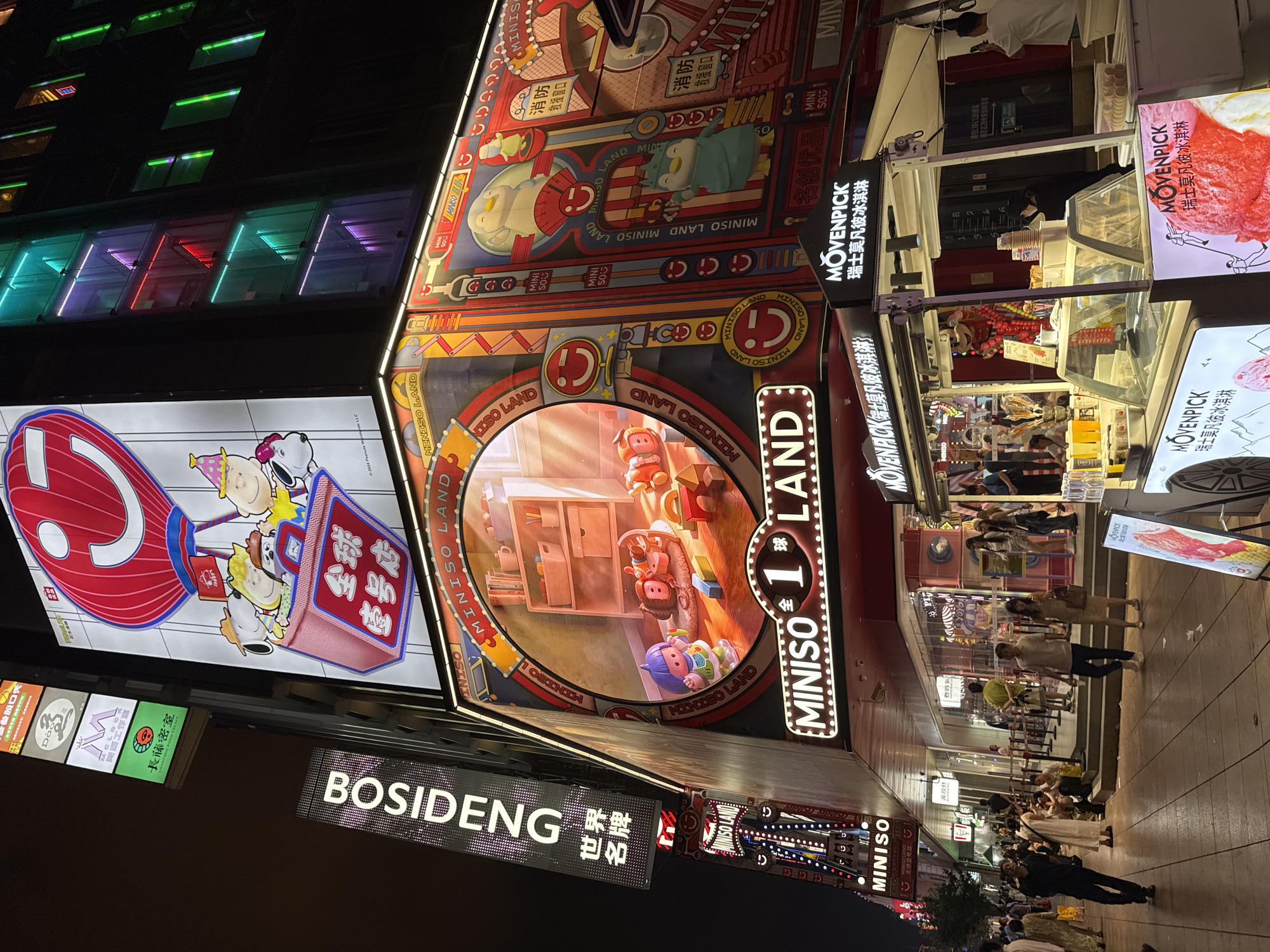 The image depicts a street scene at night in Shanghai, China, specifically on Nanjing Road, a well-known shopping street. The foreground features a Movenpick ice cream stand with several people browsing and purchasing items. The Movenpick branding is prominently displayed in English and Chinese. Behind the ice cream stand, the façade of a multi-story building is covered in large, brightly lit advertisements.
One of the most prominent advertisements is for "MINISO LAND," a store that appears to be a toy or novelty shop. This advertisement features animated characters, including Snoopy and other Peanuts characters, riding in a hot air balloon, as well as a circular display showcasing toys in a room setting. Another vertical sign reads "BOSIDENG," a Chinese down jacket brand, with smaller Chinese characters below it that translate to "World Famous Brand." The building itself is illuminated with colorful LED lights, primarily green and red, accentuating its modern architecture.
The street is paved, and there are multiple shops and storefronts visible, some with their entrances open and customers inside. Several individuals are walking along the sidewalk, while others are gathered around the shops or the ice cream stand. The overall atmosphere is vibrant and bustling, characteristic of a busy urban shopping district at night. The lighting creates a dynamic and somewhat surreal ambiance, with the bright advertisements casting a glow on the street and the surrounding buildings.