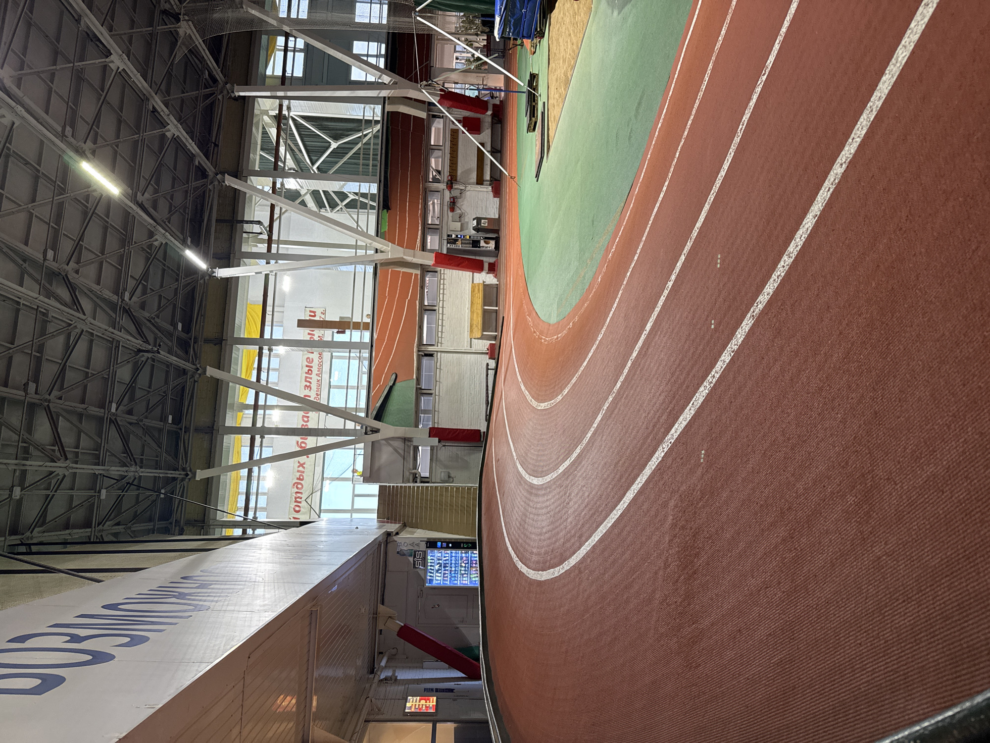 This image depicts the interior of a modern indoor athletics track facility. The track itself is a vibrant red, with white lane markings clearly visible. It curves prominently in the foreground, leading into the mid-ground. Adjacent to the track, a green surface, likely for field events, is visible. In the background, a complex metal ceiling structure with lighting fixtures supports the roof of the building. White support pillars are strategically placed throughout the space. Various elements of the facility are visible, including a vending machine stocked with beverages, stairs leading to an upper level, benches, and what appears to be a sandpit for jumping events. Large banners with Russian text are hung on the walls, suggesting this is a sports complex or stadium. The overall environment is well-lit, clean, and equipped for athletic training and competition.