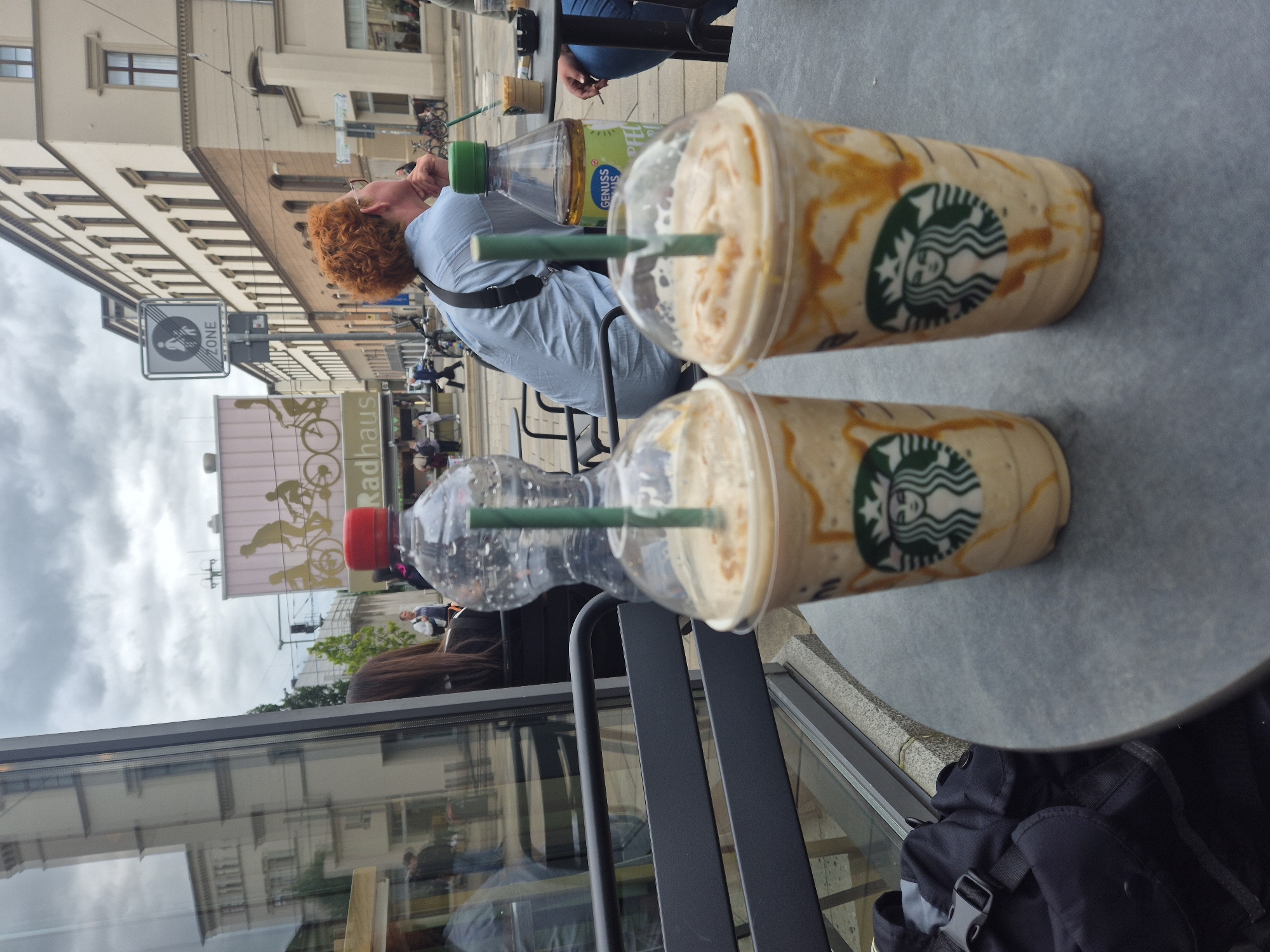 The image captures an outdoor café scene, primarily focusing on a gray tabletop where two Starbucks Frappuccino drinks with green straws are placed. Alongside them are two clear plastic bottles: one with a green cap and the visible text "GENUSS NATUR," and another with a red cap.
A person with curly red hair, wearing a light blue collared shirt and glasses, is seated at the table, resting their head on their hand, facing towards the street. The back of another person's head with long dark hair is partially visible, seated opposite the red-haired individual. A black backpack is on the ground next to the table.
The background reveals a bustling city street with multi-story buildings featuring multiple windows and neutral-colored facades, suggesting European architecture. The sky is overcast with visible clouds. Several street signs are present, including one clearly marked "ZONE" with a pedestrian icon. A prominent banner on a building in the mid-ground displays imagery of bicycles and the German word "Radhaus," indicating a bicycle-related business or area. Reflections of the surroundings are visible in a large window adjacent to the outdoor seating. The scene depicts a casual daytime moment, likely in the late morning or afternoon, at an urban cafe.