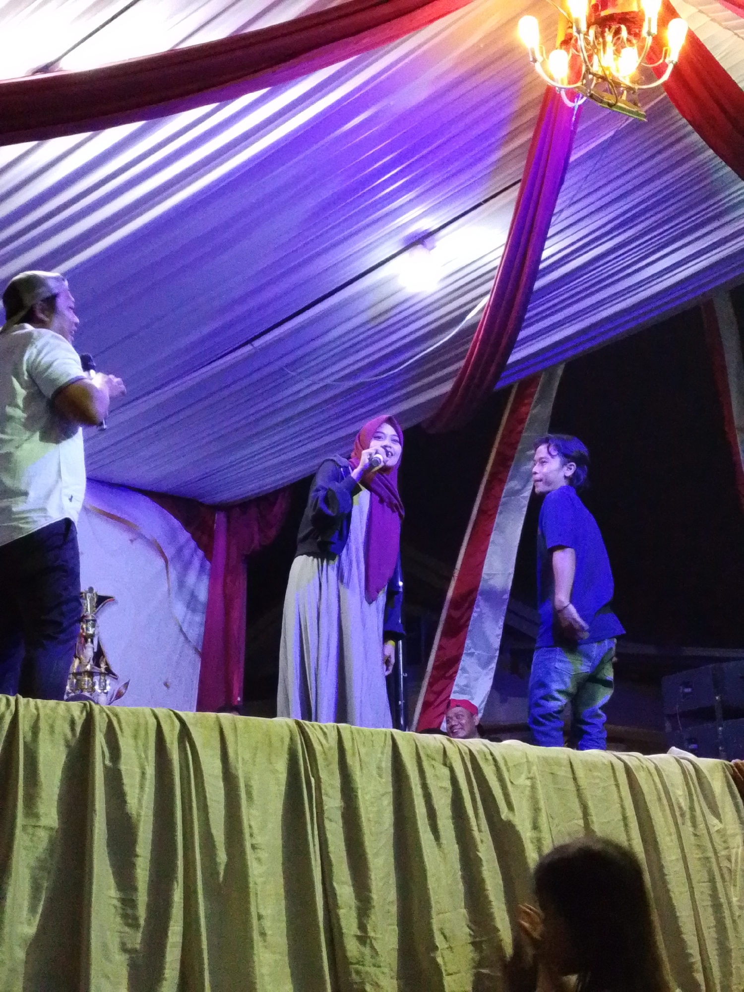 This image depicts an outdoor event at night, possibly a performance or gathering, in Lawatan, Indonesia. A stage is set up with a backdrop of draped fabric, illuminated by purple and white lights, creating a festive atmosphere. In the foreground, a man in a white shirt and a baseball cap is holding a microphone, appearing to speak or sing. To his right, a woman in a hijab and a long grey dress is also holding a microphone, seemingly engaged in the same activity. A young boy in a blue shirt and jeans stands to the right of the woman, looking towards the performers. The stage is covered with a green fabric, and in the background, there are large speakers and a decorative chandelier with multiple light bulbs. A person's head is visible peeking over the front edge of the stage, and another person's silhouette is present in the lower right corner, looking towards the stage. The overall scene suggests a community event or cultural performance taking place in Lawatan.