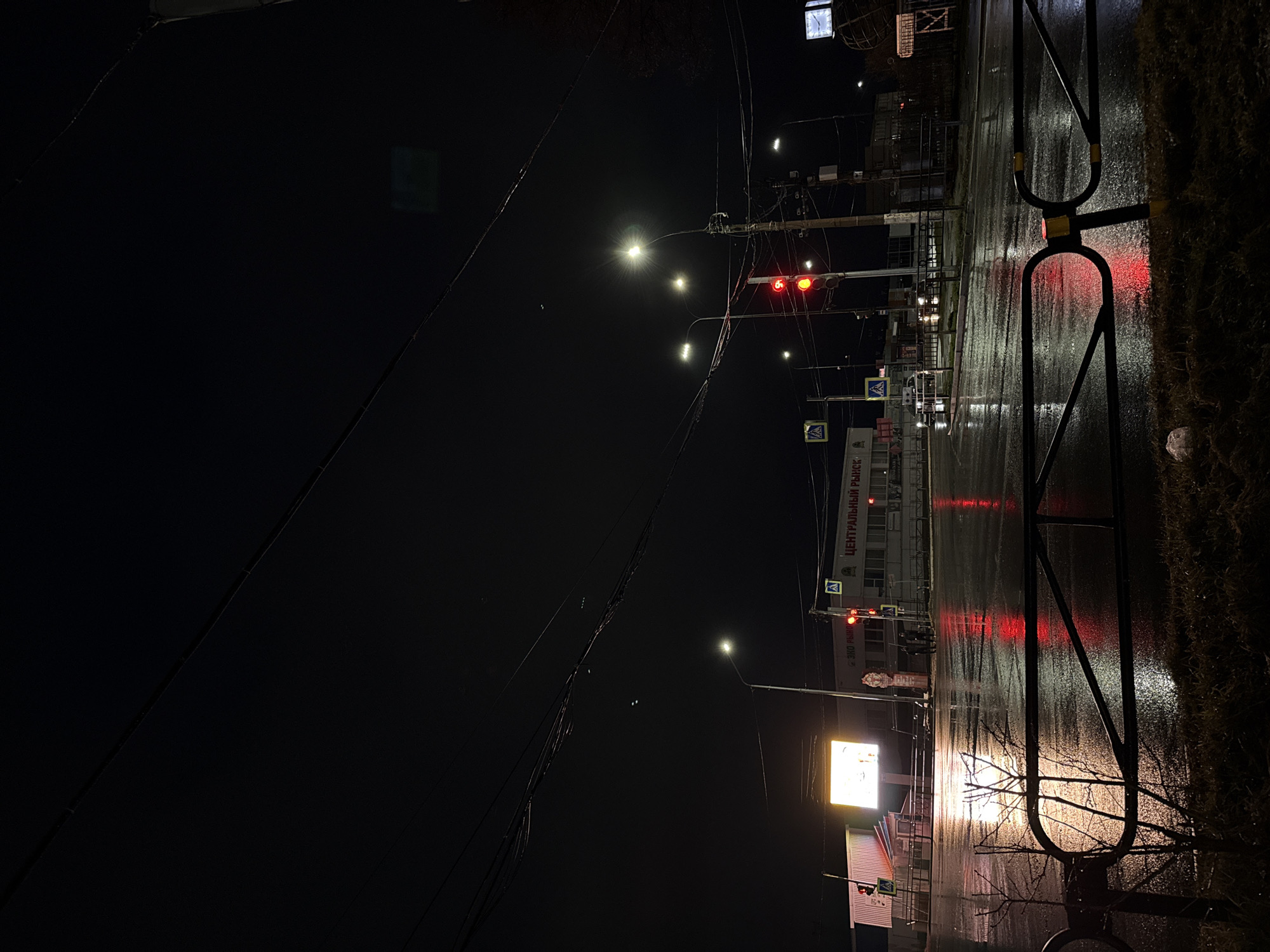 The image depicts a nighttime street scene in Chaikovsky, Russia, possibly after a rain shower, indicated by the wet pavement reflecting light. A crosswalk barrier is visible on the right edge of the frame. A building, identified by the text "Центральный рынок" (Central Market) in Russian, is visible in the background. Traffic lights are present, displaying red. Streetlights illuminate the scene. Overhead power lines are present. A billboard with a bright display is seen on the left side of the frame. Pedestrian crossing signs are visible. The scene appears to be a regular urban environment at night.