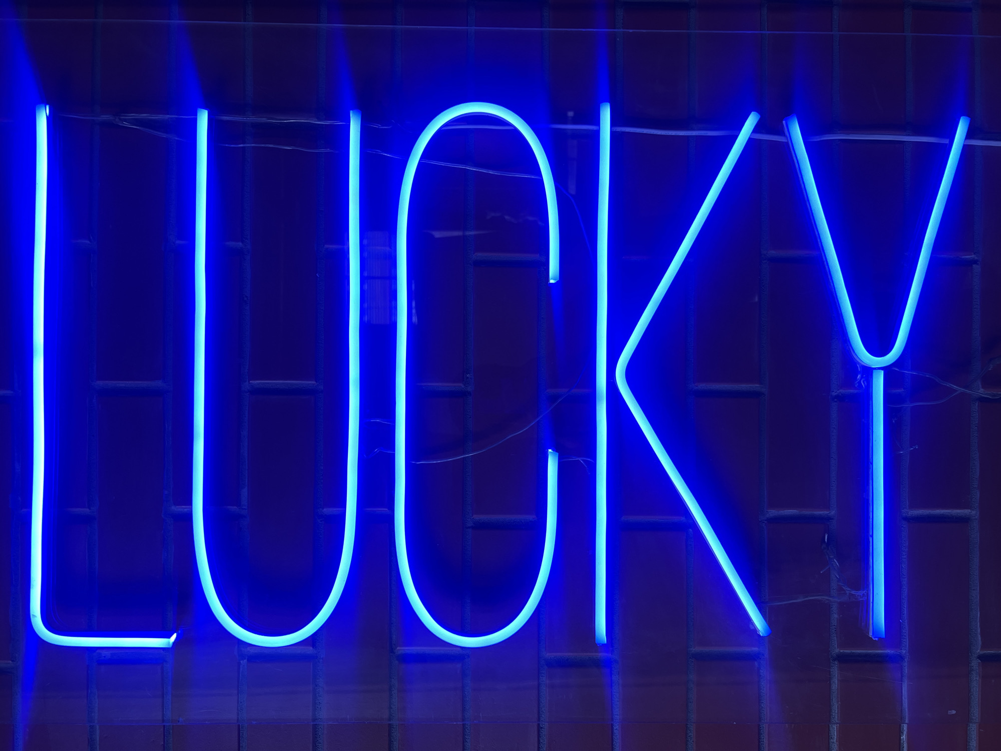 The image shows a neon sign on a brick wall. The sign is made of blue neon tubes and spells out the word "COOK". The brick wall is red and has a textured surface. The lighting is dim, with the neon sign being the primary source of illumination. The image does not provide any specific information about its location in Moscow, Russia. There are no people or any other objects visible in the frame besides the neon sign and the brick wall. The overall impression is one of a stylized or artistic display.