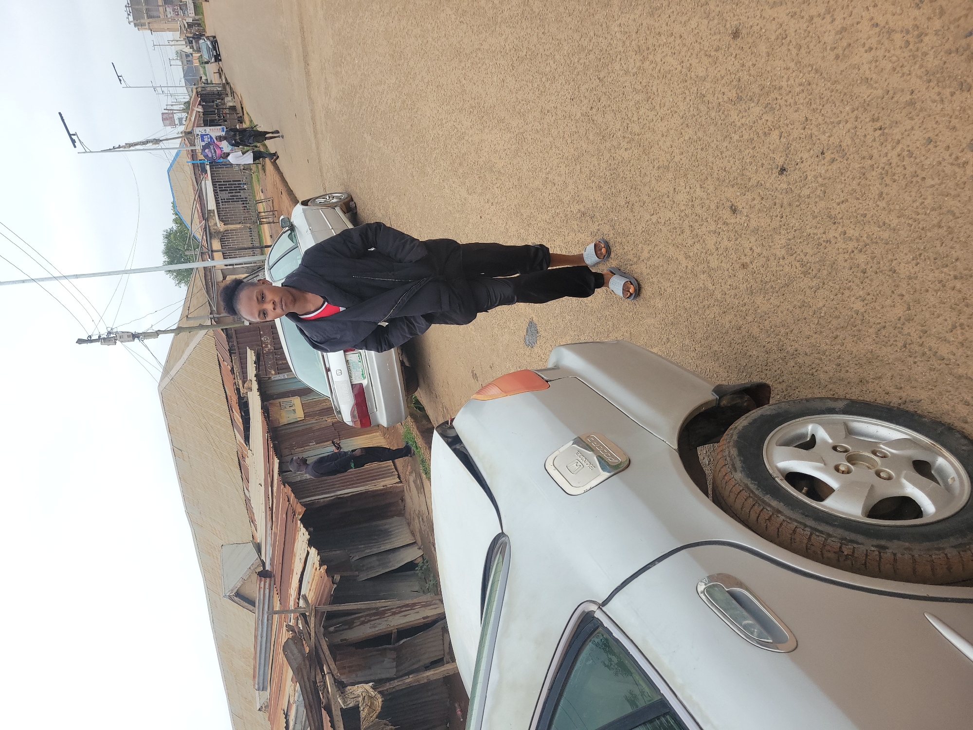 A young woman with her hair pulled back in a bun stands on a dirt road in front of a silver Honda Accord. She is wearing a black hooded jacket and black pants, with a red shirt visible underneath her jacket. She has on grey slide sandals. The car's trunk is open and bent. On the left side of the image, there are several corrugated metal buildings. In the background, there are more buildings, streetlights, and electrical poles. Two other people are visible walking down the street in the background, and another car is parked further down the road. The sky is overcast.