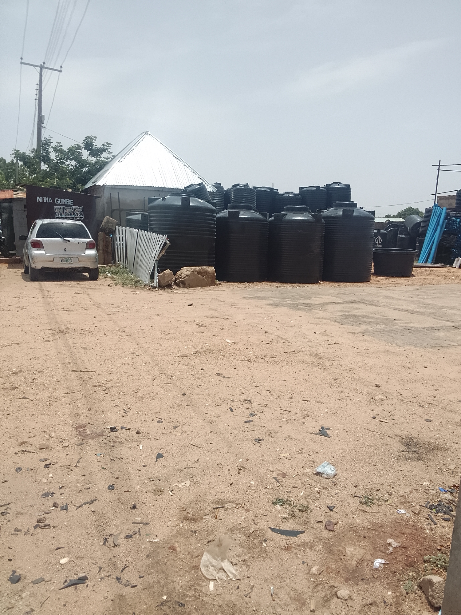 The image depicts an open-air commercial space in Pantame, Nigeria. A collection of large, black plastic water storage tanks occupies the center of the frame, arranged in rows. In the background is a building with a pyramidal, corrugated metal roof. To the left, a silver car is parked, its rear facing the camera. The foreground is a dirt ground scattered with debris and small stones. A sign reading "NINA GOMBE" is visible above the parked car. A utility pole is visible on the left side of the image.