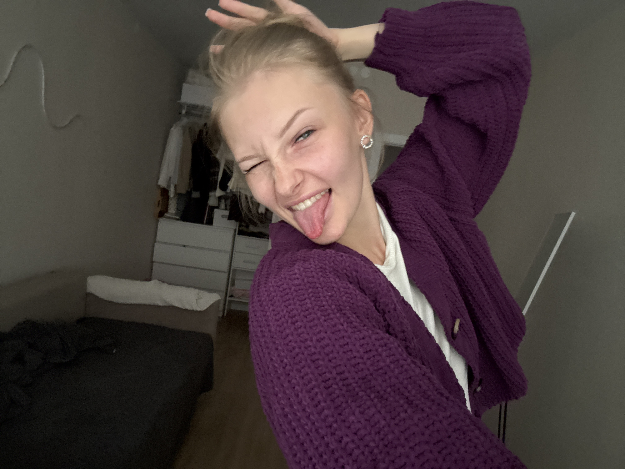 A young Caucasian female is depicted in a selfie taken in a bedroom. She is wearing a purple knitted cardigan over a white shirt and is playfully sticking her tongue out, winking, and has her hand on her head. The room contains a bed, a dresser, a closet with clothes, and a mirror. The lighting is dim, suggesting it might be evening. The location is identified as Perm, Russia.