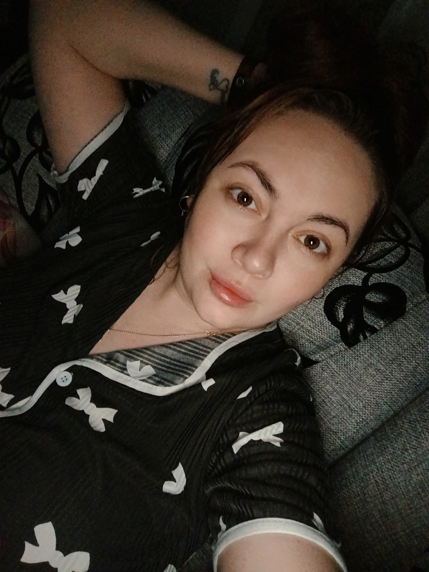 The image features a woman lying down indoors, likely on a couch or bed, in Chaikovsky, Russia. She is captured in a close-up, looking directly at the camera with a neutral expression and slightly parted lips. Her fair skin, brown eyes, and dark hair are prominent. She is wearing a dark, possibly black, shirt or pajama top adorned with white bow patterns and white piping along the collar and placket, which includes visible light-colored buttons. A delicate gold chain necklace is visible around her neck, and a small hoop earring is in her right ear. A tattoo is discernible on her left arm, which is bent and positioned behind her head. The lighting is dim, suggesting an evening or nighttime setting. The background is composed of a textured gray fabric, consistent with upholstery from a sofa or cushions. No visible text is present in the image.