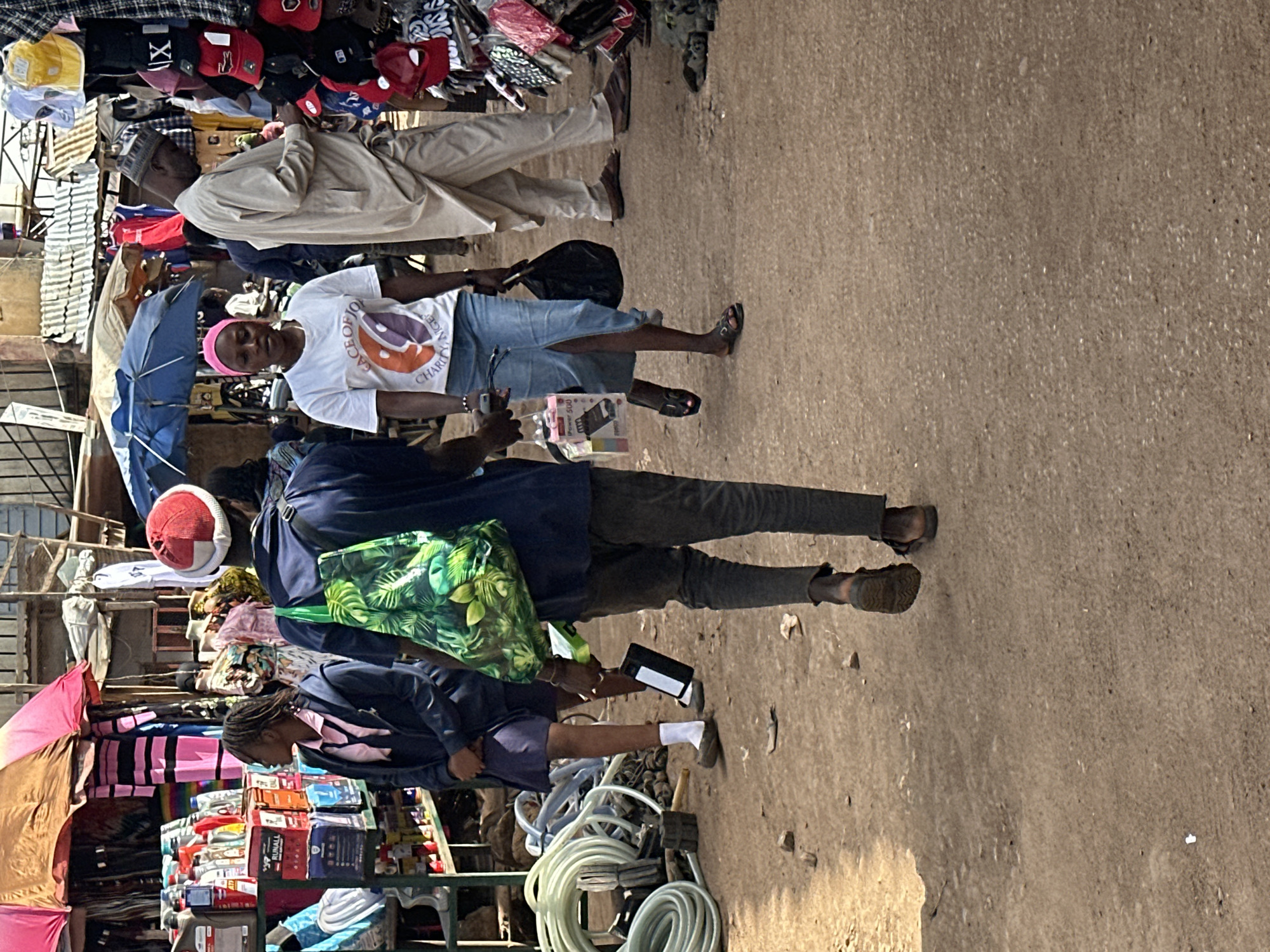 The image depicts a bustling outdoor market scene in Bukuru, Nigeria. In the foreground, a person walks away from the camera, carrying a green patterned bag and a dark object. To their left, a young person in a school uniform, consisting of a dark jacket, pink shirt, and blue skirt, stands facing left, looking at the market stalls. To the right of the foreground figure, a woman with a pink headscarf and a white t-shirt with a graphic print walks towards the left, carrying a black bag. Behind her, a man in a light-colored traditional outfit stands, looking down at something in his hands. The market stalls are filled with various goods, including clothing, hats, and what appear to be bottles of liquids. The ground is dirt and gravel, and umbrellas are visible providing shade over some of the stalls. The environment suggests a lively commercial area with multiple people engaged in shopping and selling.