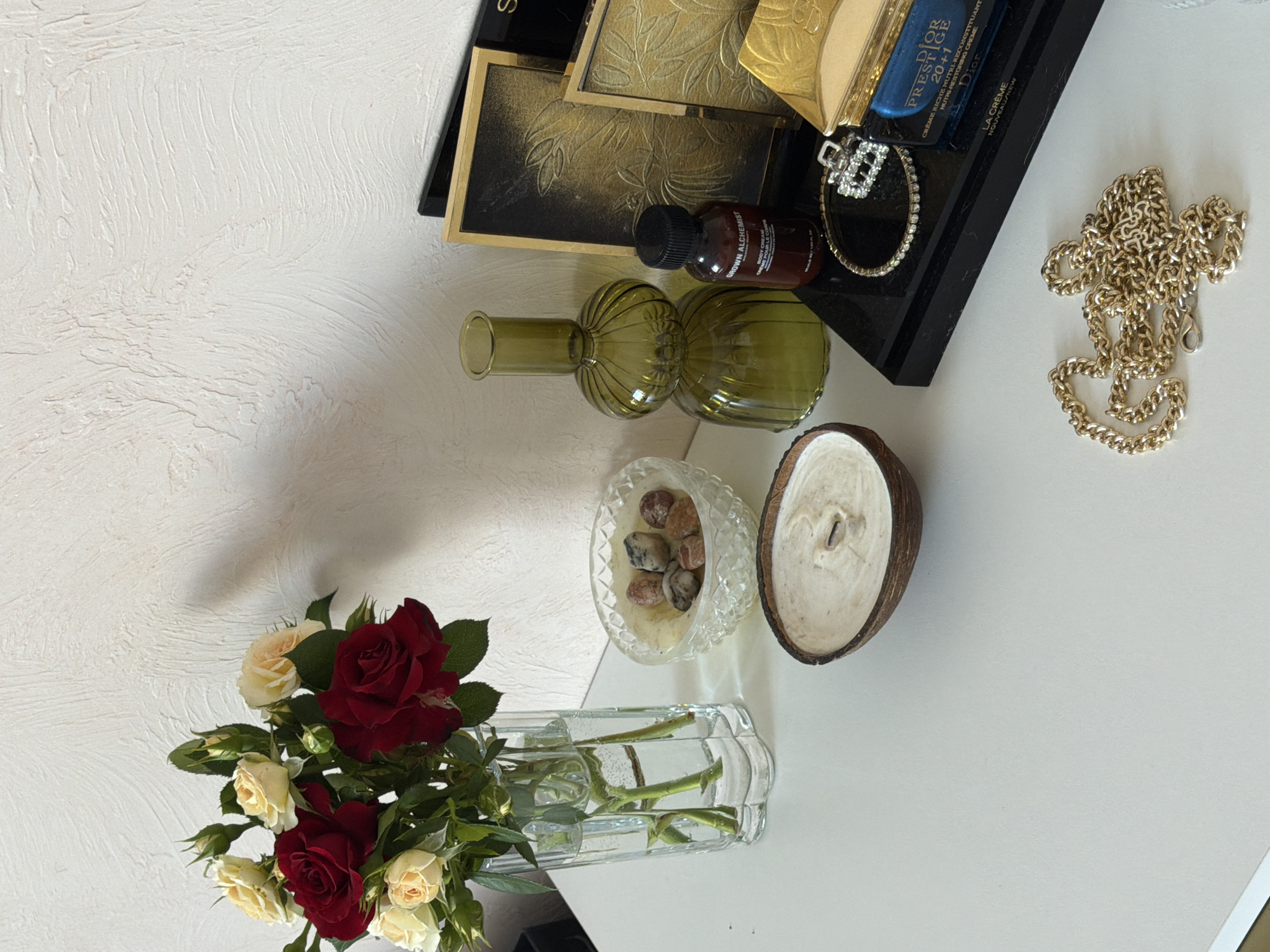 A still life arrangement is displayed on a white surface against a textured white wall. In the upper left corner, a bouquet of red and cream roses in various stages of bloom is held in a clear glass vase filled with water. To the right of the vase is a clear, faceted glass bowl containing a cream-colored substance topped with a cluster of small, smooth stones. In front of the bowl is a coconut shell half filled with a white, waxy substance, presumably a candle, with a visible wick.
Further to the right, a dark green glass vase with a rounded, segmented design stands next to a small brown bottle labeled "BROWN ALCHEMIST BODY CREAM". Behind these items are two framed objects with ornate gold leaf designs on a dark background.
In the foreground, a black tray holds several items. A delicate silver bracelet with clear stones is positioned on the tray, next to a small, gold-colored hinged box. Below the gold box is a blue and silver product labeled "DIOR PRESTIGE 20+1 CRÈME RICHE NUTRITIVE RESTORING CREAM". A gold chain necklace is draped loosely on the white surface in the lower right portion of the image.
The scene appears to be an indoor still life arrangement, with no indication of its specific location within Rossiyskiy, Russia, or any actions or interactions taking place.