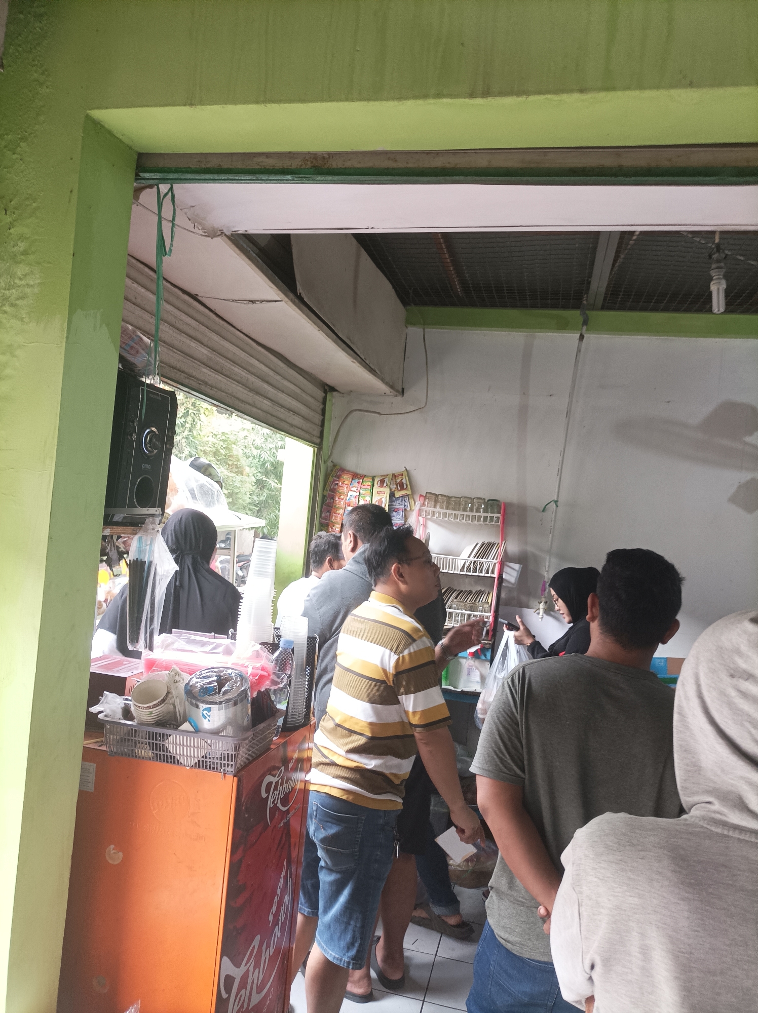 The image shows a busy outdoor food stall in Cirebon, Indonesia. Several people are queuing to order or pick up food. A woman in a black headscarf is visible behind the counter, possibly serving customers. In the foreground, a man in a striped shirt and denim shorts is talking to someone. The stall appears to be selling drinks, with stacks of plastic cups and bottles on display. The overall environment is casual and bustling, typical of a street food establishment.