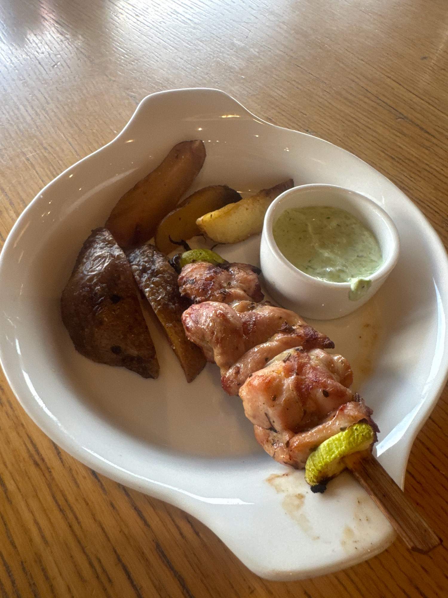 A white plate holds a skewer of grilled chicken with zucchini and potato wedges. A small bowl of green sauce sits to the side. The plate rests on a wooden table. This image was taken in Irkutsk, Russia.