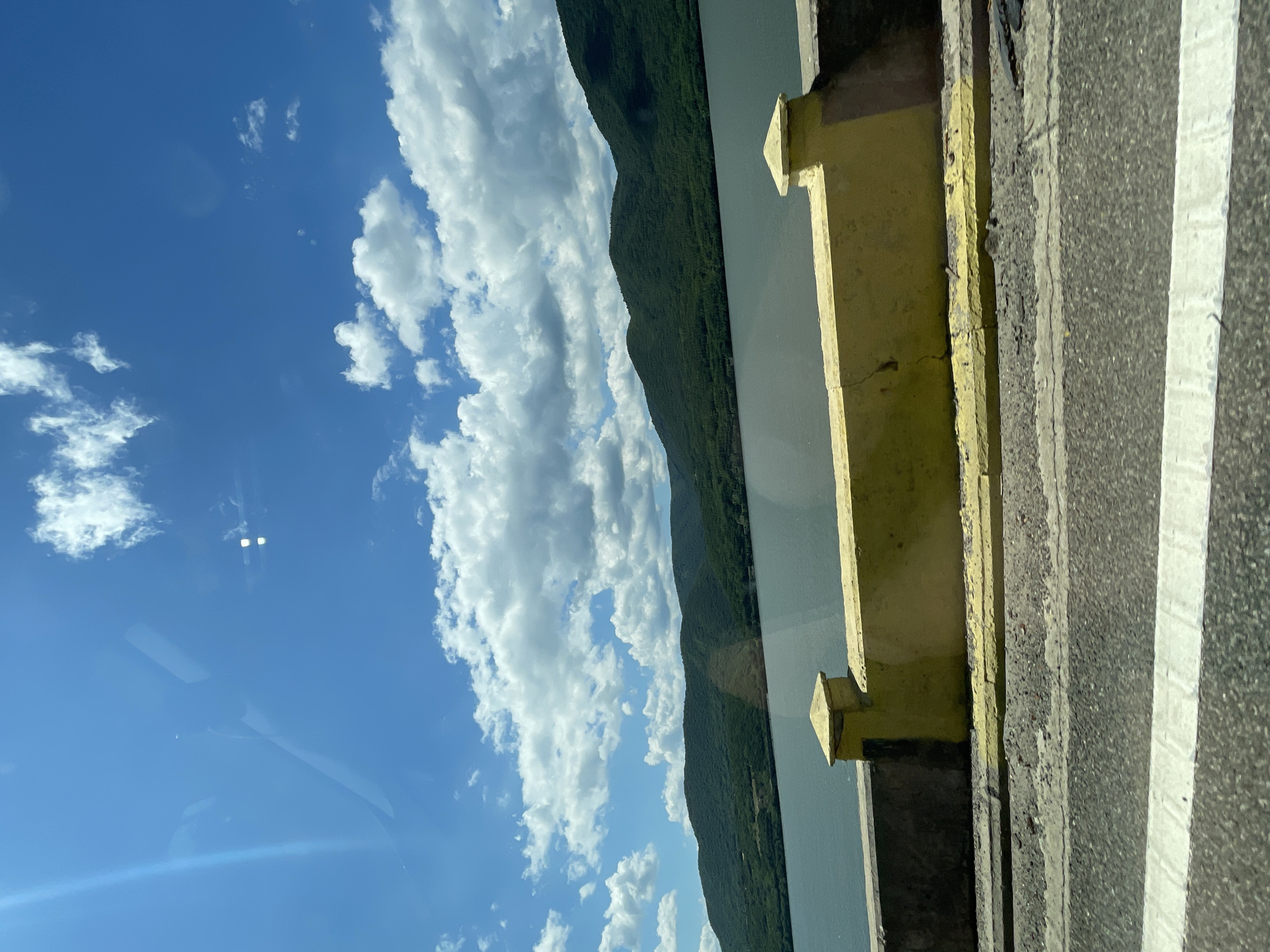 The image captures a scenic outdoor landscape in Drobeta-Turnu Severin, Romania, viewed through a window, likely from a moving vehicle. The scene is dominated by a clear blue sky with numerous scattered white cumulus clouds. The upper left portion of the sky exhibits a prominent diagonal light flare or reflection, along with indistinct lighter shapes, suggesting reflections from the interior of a vehicle. Two distinct, bright, small rectangular reflections are visible in the upper right sky area.
Below the sky, a vast body of dark blue-grey water extends horizontally across the mid-ground. Beyond the water, a series of lush green, densely forested mountains or hills rise, forming a continuous undulating horizon line across the distant background.
In the foreground, a concrete barrier is visible. The primary section of the barrier is painted a bright yellow, with a small square, yellow-capped post-like structure positioned towards its left. To the immediate left of this post, the barrier appears to be unpainted grey concrete. Below the main yellow barrier, a narrower yellow curb or ledge runs parallel. The very bottom of the image shows a section of dark asphalt road, featuring a thick, solid white line running horizontally, indicative of a lane marker. No people or animals are visible in the scene, and no specific actions are occurring. The environment suggests a sunny day with good visibility, highlighting the natural beauty of the region alongside man-made infrastructure.