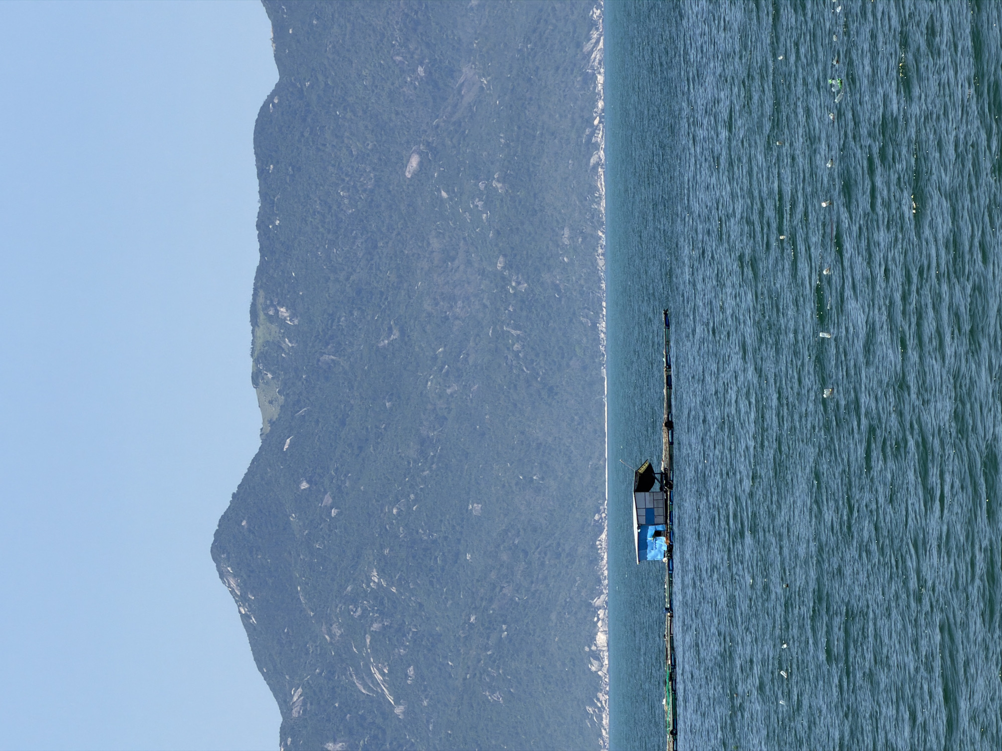The image captures a coastal scene in Vĩnh Lương, Vietnam, under a clear, light blue sky. A sprawling mountain range, covered in dense green vegetation interspersed with large rock formations, stretches across the background, exhibiting a blue-grey hue due to atmospheric distance. The lower two-thirds of the frame are occupied by the deep blue-green, rippling surface of the sea. In the mid-ground, a small, weathered structure with a blue side and white roof floats on a series of interconnected rafts or pontoons, part of a larger aquaculture or fishing platform. Scattered across the water's surface in the foreground and mid-ground are multiple pieces of plastic litter, predominantly white and green bottles. No people or animals are visible.