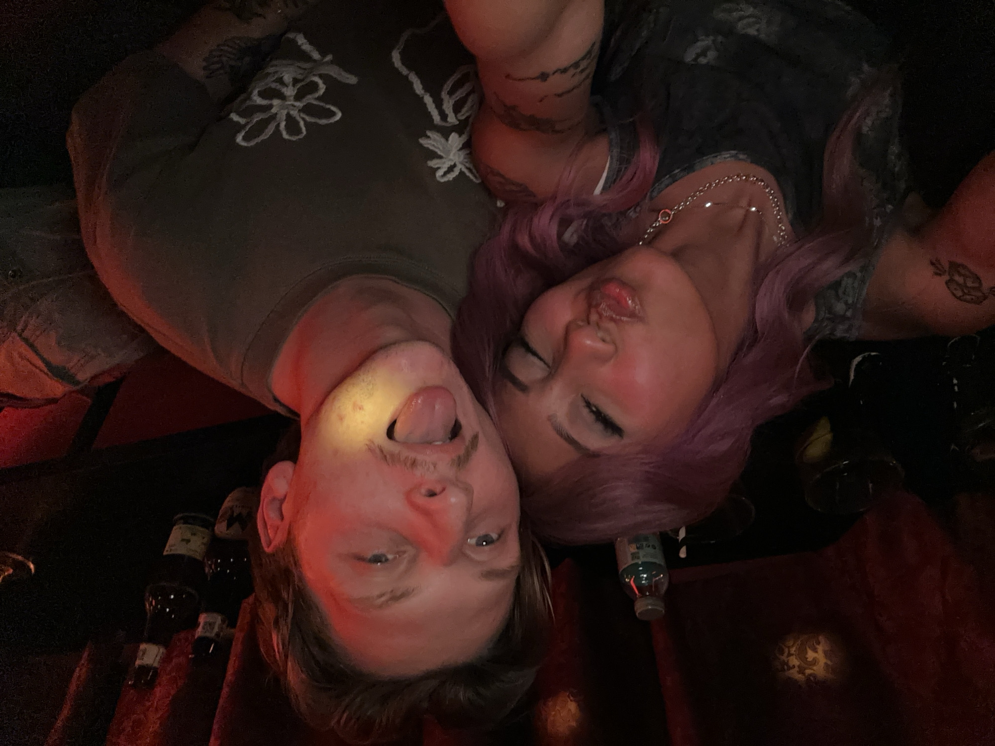 Two people are posing for a photograph. The man on the left has fair skin, short hair, and a mustache; he is sticking his tongue out. The woman on the right has pink hair and is pursing her lips. They are both wearing casual clothing and have tattoos on their arms. There are multiple bottles and glasses in the dark background, as well as red velvet drapery. A water bottle is on the table. It is impossible to identify the precise location of this scene, or even confirm it to be in Barcelona, Spain, without further information.