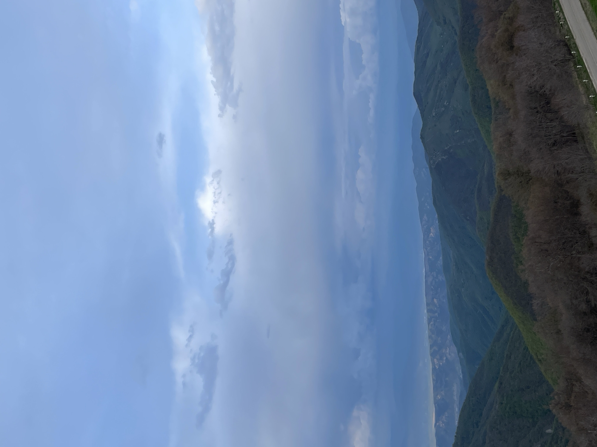 The image depicts a mountainous landscape under a cloudy sky in Zemo Nichbisi, Georgia. A range of green mountains occupies the right side of the frame, with a glimpse of a road on the far right edge. The sky, predominantly filled with light blue and grey clouds, takes up the majority of the image on the left side. The scene is shot from an elevated perspective, capturing the scale of the mountains and the vastness of the sky.