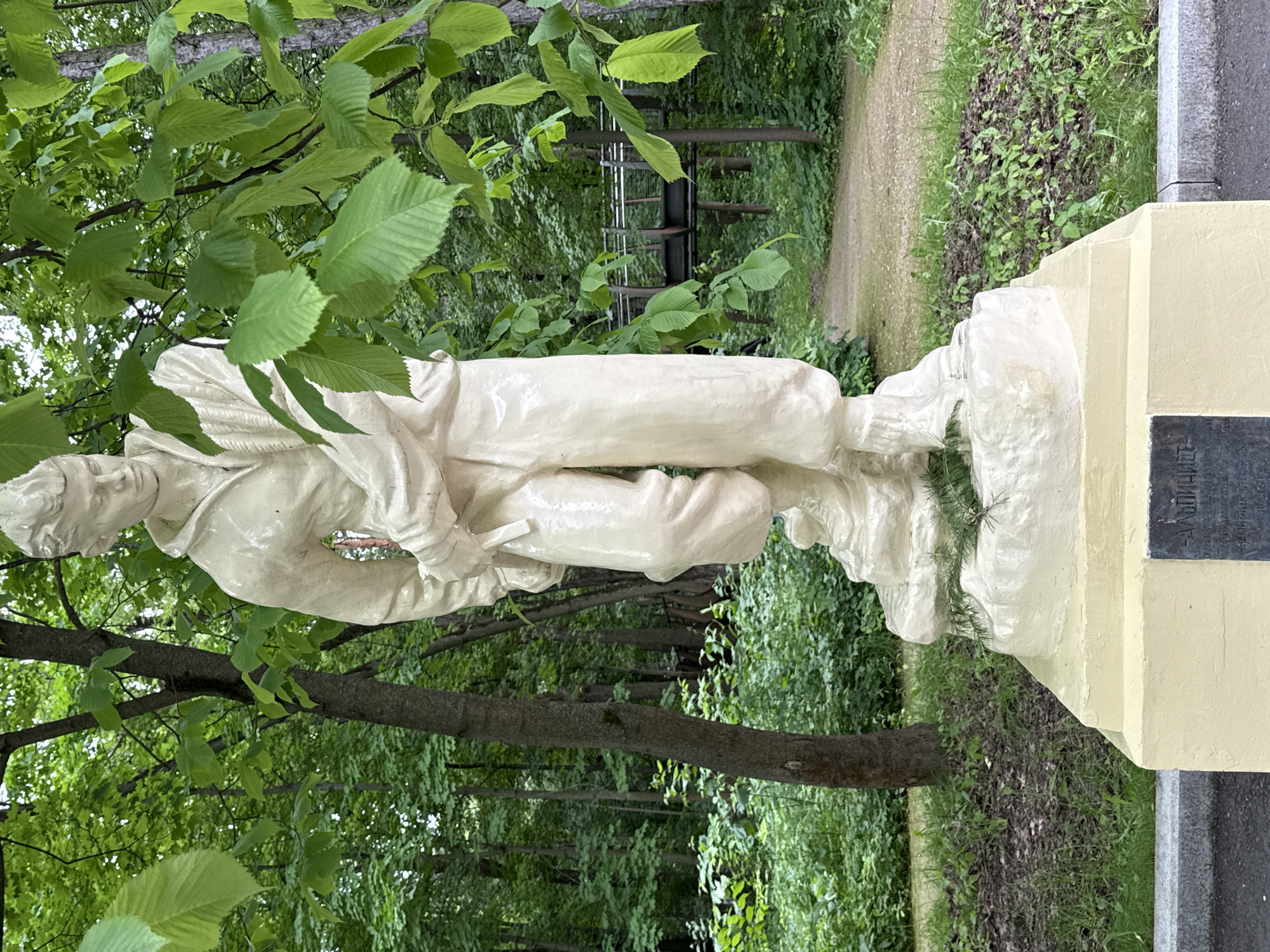 The image shows a light-colored statue of a man in outdoor clothing standing on a pedestal in a park. The statue is a full-body representation, with the man appearing to be in his late teens or early twenties. He is looking down and to his right, with his arms crossed in front of him. The statue is made of a smooth material, possibly plaster or concrete, and has a matte finish.
The statue is located in a park with mature trees and lush green foliage. There is a walking path behind the statue, and a pedestrian bridge can be seen in the background. The weather appears to be fair, with sunlight filtering through the leaves of the trees.
The inscription on the plaque at the base of the statue is in Russian and reads "СЛАВНЫЕ РАБОЧИЕ" which translates to "GLORIOUS WORKERS". This suggests the statue is a monument to laborers. The statue is located in Moscow, Russia, specifically in the Sokolniki Park. The overall impression is one of remembrance and honor towards the working class.