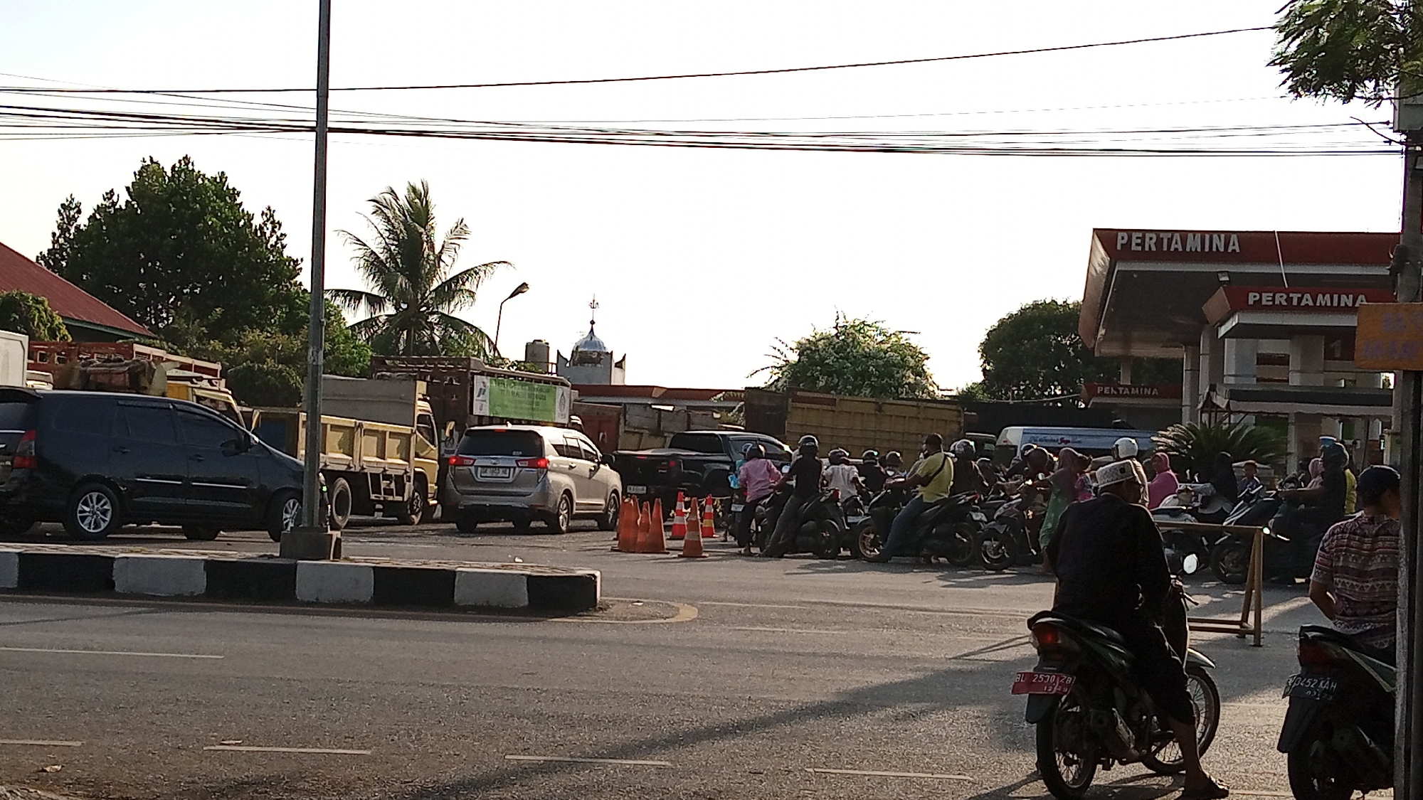 The image captures a street scene in Julikeude Dua, Indonesia, likely a busy intersection or near a gas station. In the foreground, a paved road with a concrete divider marked with black and white stripes is visible. Several orange traffic cones are placed on the road, suggesting temporary traffic control or an event.
A variety of vehicles are present. A black SUV is parked on the left, and a silver SUV is further down the road. Multiple motorcycles are lined up, some with riders, indicating a queue or waiting period. Several trucks, including one with a yellow bed, are also part of the traffic.
In the background, a Pertamina gas station with its distinctive red and white signage is prominent. Behind the gas station, trees and buildings, including a structure with a dome that resembles a mosque, are visible. The sky is bright and clear, suggesting daytime.
People are seen on and around the motorcycles, some wearing traditional headwear. Their actions suggest they are either waiting for fuel, a ride, or observing the surroundings. The overall atmosphere appears to be one of a normal, bustling day in a populated area of Julikeude Dua.