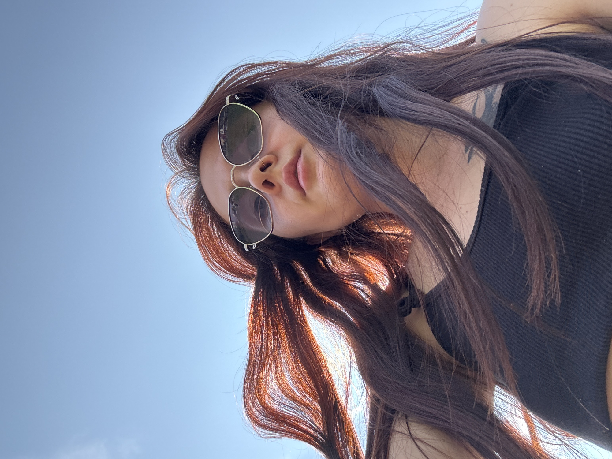 A low-angle, outdoor, waist-up shot captures a young woman looking up towards the camera with her long, dark auburn hair blowing in the wind against a clear blue sky. She is wearing sunglasses with thin metal frames and a black ribbed tank top. A tattoo is visible on her left shoulder. The lighting suggests it is daytime, possibly sunny. The image's perspective and the clear sky imply the woman is outdoors. The context provided suggests this photograph was taken in the city of Naberezhnye Chelny, Russia.