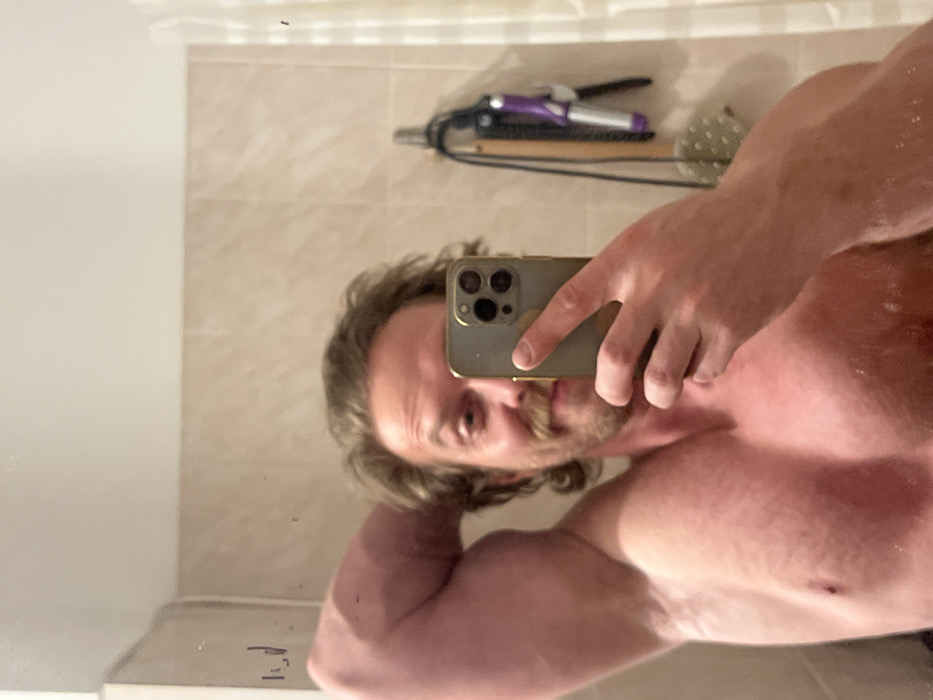 A mirror selfie captures a torso, upper arm, and head with a mustache, light brown hair, and beard. A phone is held in front of his face, obscuring part of it. The background reveals a beige tiled wall, a curling iron, and a hairbrush. The lighting is soft, creating a slightly blurry reflection.