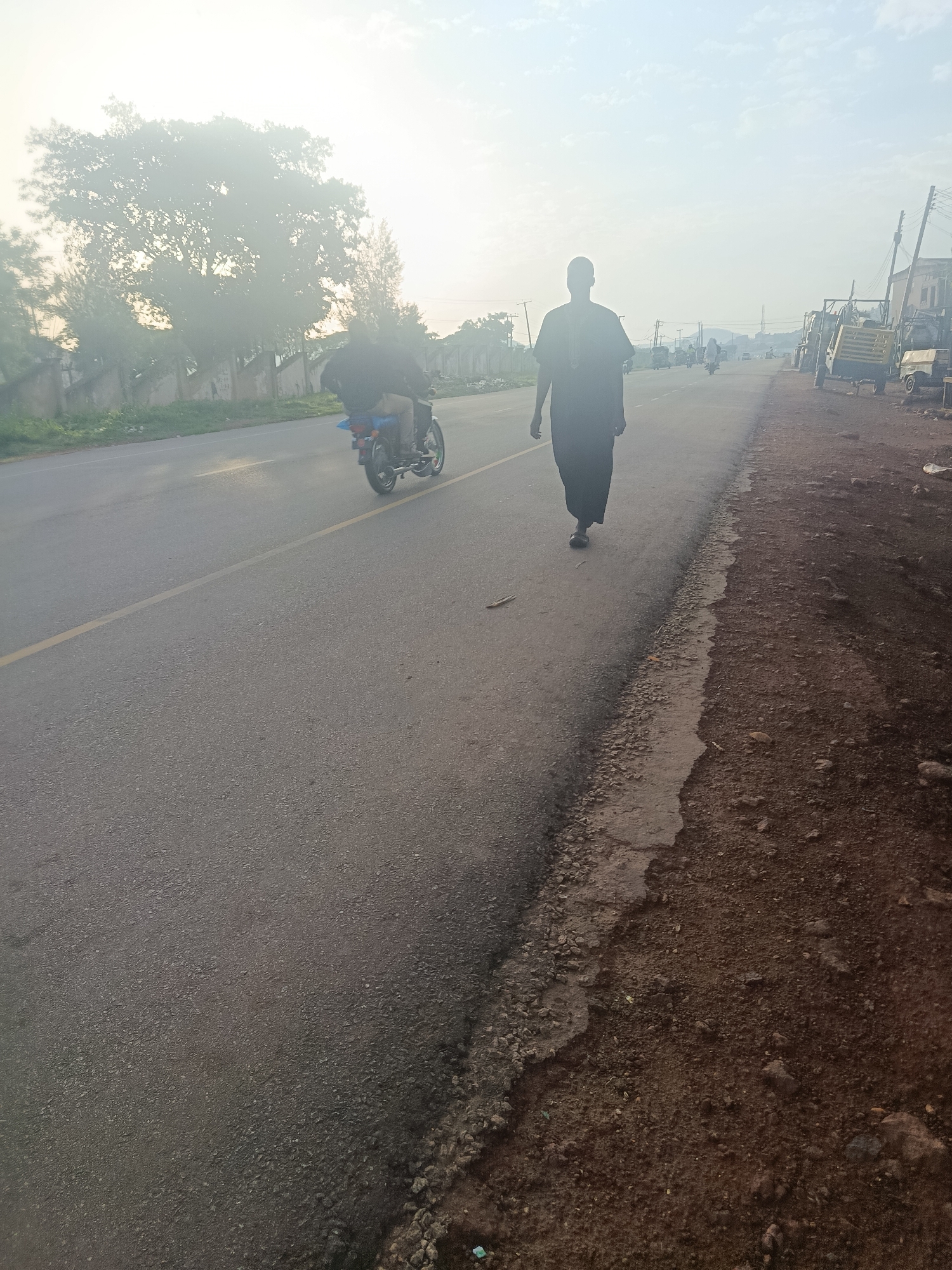 A man in dark traditional attire walks along the shoulder of a paved road, facing away from the camera. The road has a yellow line marking its center. To the left of the road, there are trees and a wall. To the right of the road, there is a dirt shoulder with loose rocks and debris. In the distance, several vehicles are visible on the road, including a motorcycle with a rider and several other cars and a yellow trailer. The sky is bright and hazy. The scene appears to be in a semi-urban or rural area, possibly in Rafin Par, Nigeria, given the context.