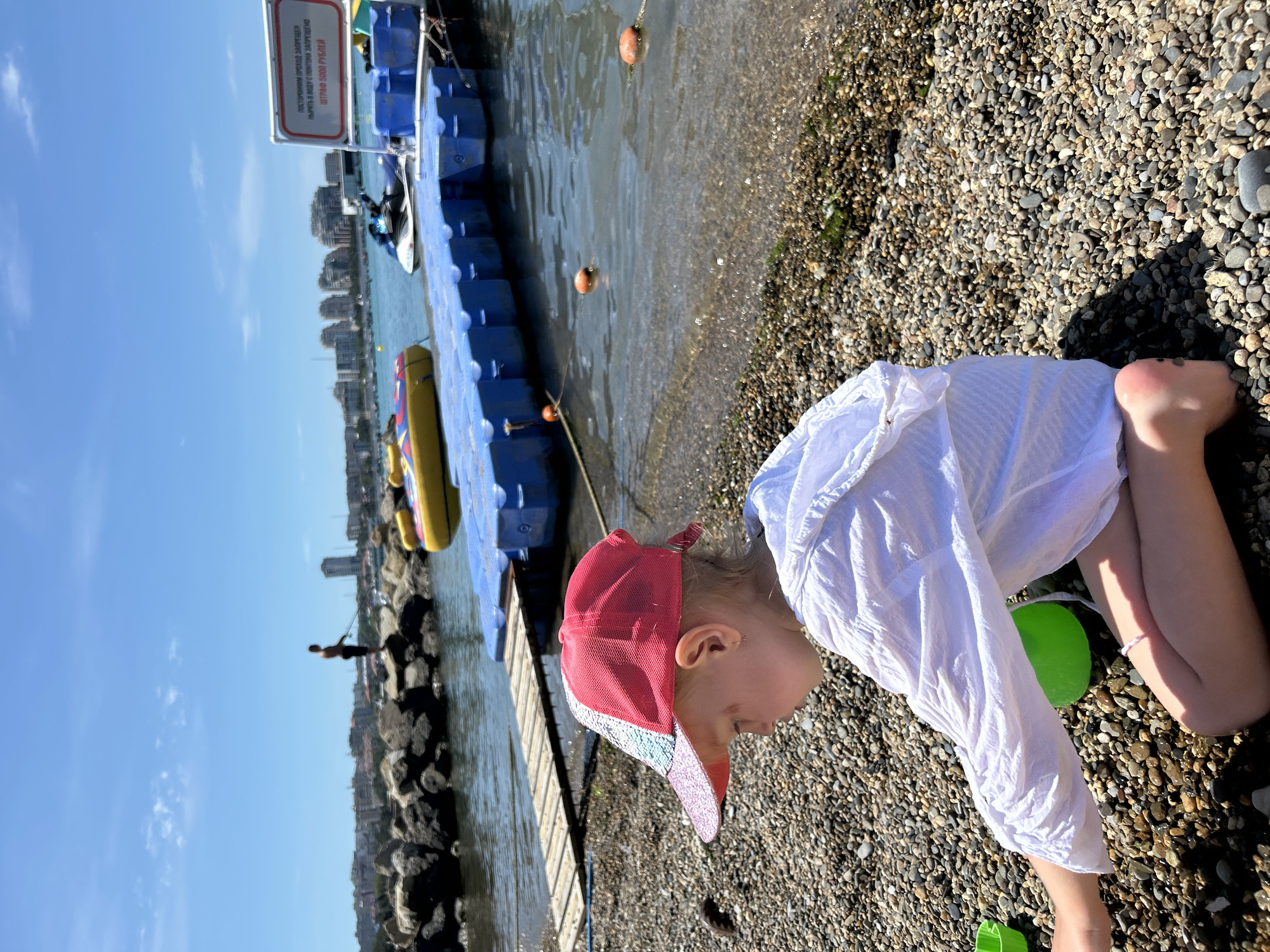 A young child, wearing a white shirt and a red and sparkly cap, is kneeling on a pebble beach. The child is facing right, with their head bowed, seemingly engaged with something on the ground. A bright green plastic toy is visible near the child's legs. The background shows a body of water, likely the sea, with blue floating platforms and a yellow inflatable object. In the distance, buildings line the coastline, and a person can be seen standing on a rocky breakwater. The sky is clear and blue, suggesting a sunny day. The overall scene appears to be set during daylight hours. A sign is attached to one of the blue floating platforms, with text that appears to be in Russian. The text on the sign reads "ПОСТОРОННИМ / ПРОХОД ЗАПРЕЩЕН", which translates to "STRANGERS / PASSAGE FORBIDDEN". Below this, it says "НЫРЯТЬ В ВОДУ С МОСТКА ЗАПРЕЩЕНО", meaning "DIVING INTO THE WATER FROM THE BOARDWALK IS PROHIBITED". Finally, it states "ШТРАФ 1000 РУБЛЕЙ", which translates to "FINE 1000 RUBLES". The image seems to capture a moment of relaxation or play at a waterfront location in Gelendzhik, Russia.