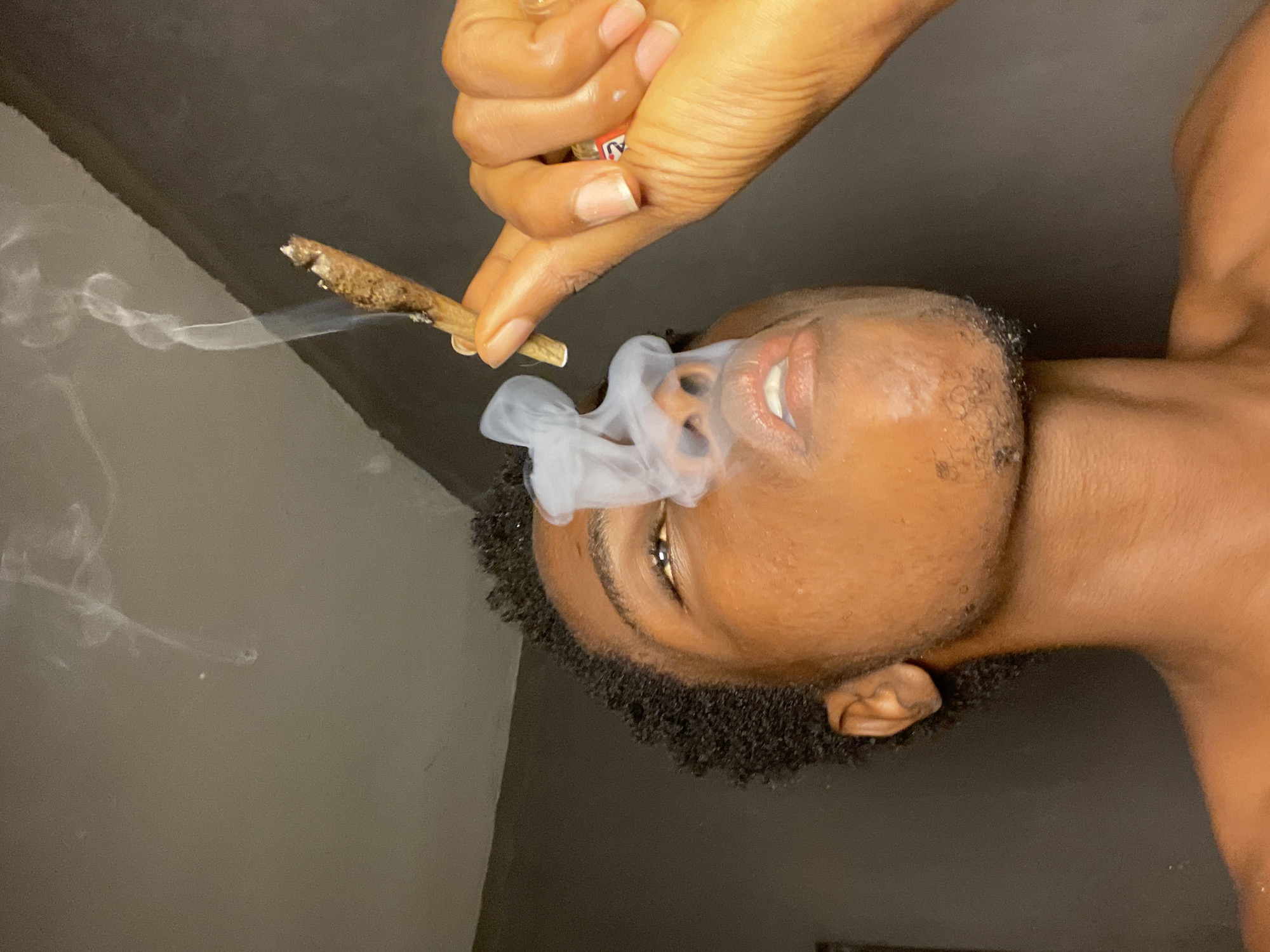 This is a close-up, low-angle shot of a young Black individual smoking a blunt. The person is holding the blunt with their right hand, and smoke is billowing upwards, obscuring part of their face. The background appears to be an indoor setting with plain, grey walls. The lighting is dim, creating shadows on the individual's face and body. The image focuses on the act of smoking, with the blunt and the rising smoke being the central elements. The scene takes place in Nkpor, Nigeria.