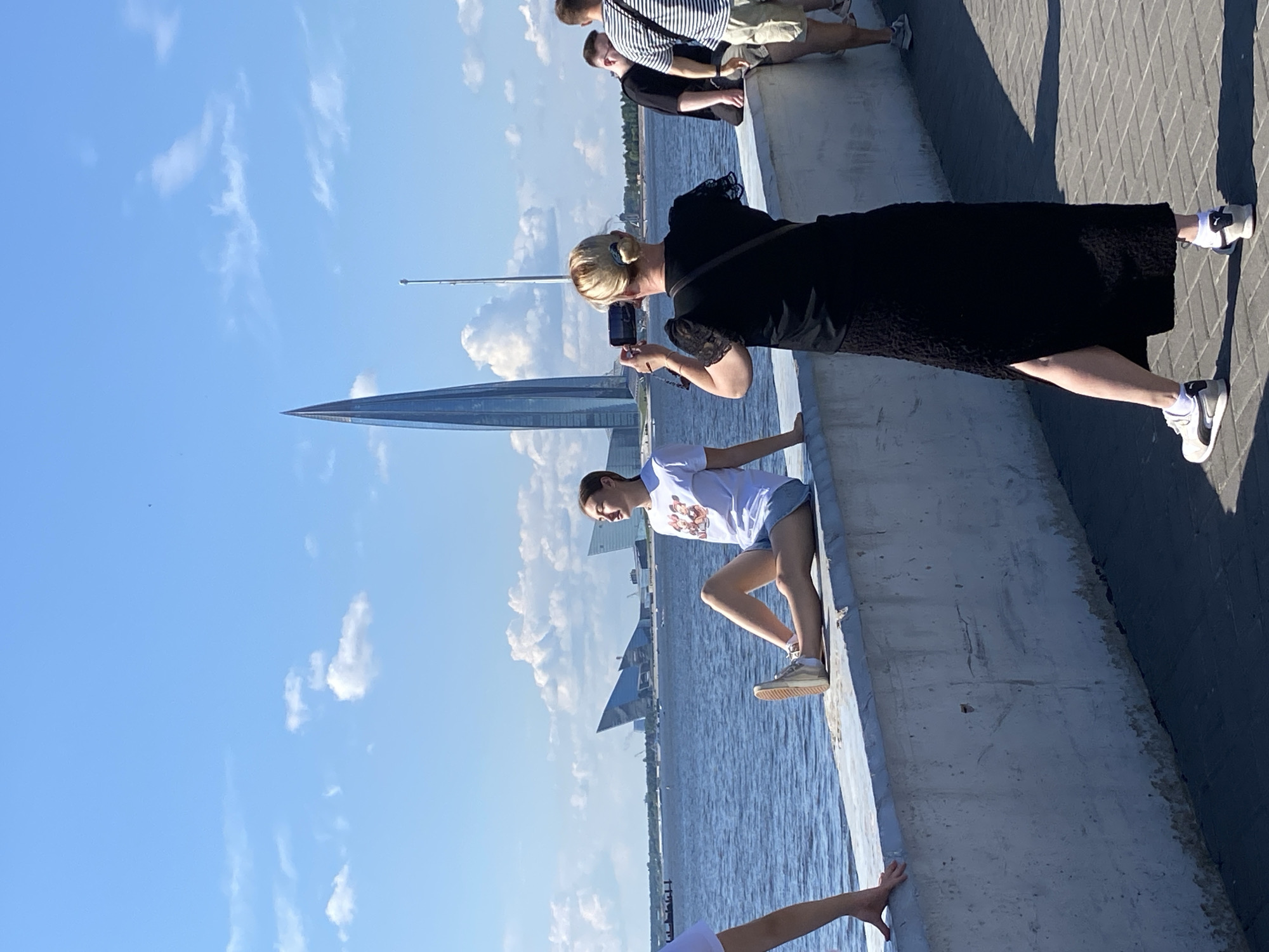 The image captures a sunny day at the St. Petersburg embankment. In the foreground, a woman in a black dress is taking a photograph with her phone of a young woman sitting on a concrete barrier overlooking the water. The young woman is wearing a white t-shirt with a graphic print and denim shorts, with her legs extended and resting on the barrier. Further to the right, two men are standing, one in a black t-shirt and the other in a striped t-shirt and shorts, also facing the water. In the background, a distinctive skyscraper with a pointed, translucent blue spire dominates the skyline, identifiable as the Lakhta Center. Several other modern buildings with angular designs are visible along the waterfront, and the Neva River stretches across the horizon under a partly cloudy blue sky. The overall scene depicts a leisurely day in a contemporary urban setting.