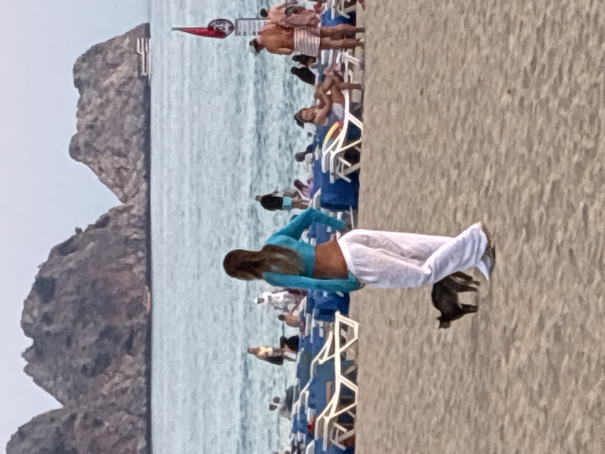 The image depicts a beach scene in Alanya, Turkey, with people lounging on beach chairs near the water. In the foreground, a woman in a teal top and white pants stands facing away from the camera, and a black cat is walking on the sand near her. In the background, there are rocky islands or cliffs in the sea, and a boat is visible near one of them. A red flag on a pole is also present. The beach is lined with blue and white lounge chairs.