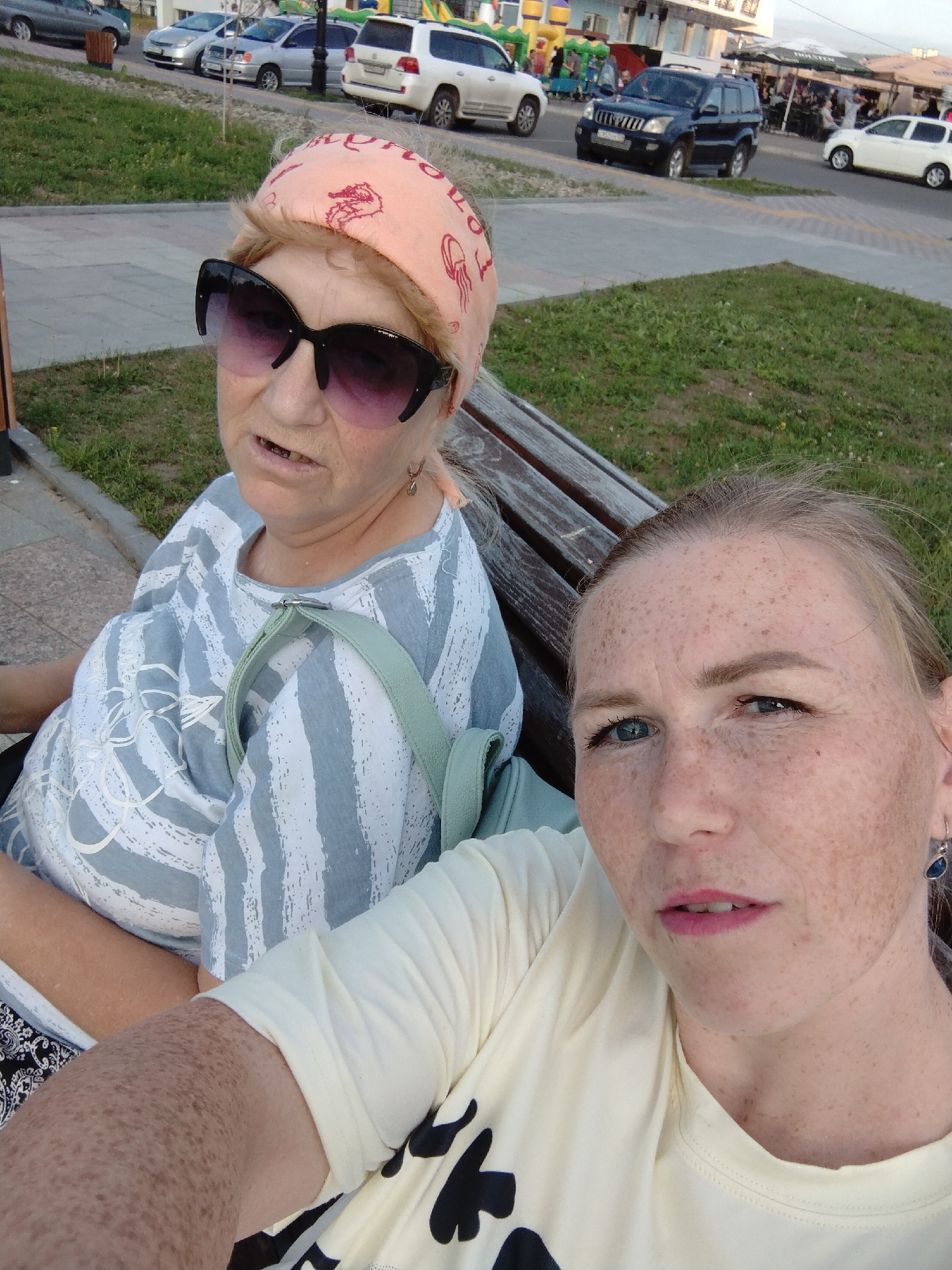 Two women are sitting on a wooden bench in an outdoor urban setting. The woman on the left is older, wearing a peach-colored bandana and large sunglasses. She has fair skin and is wearing a light blue and white striped shirt with a light green strap across her chest. The woman on the right is younger, with blonde hair and numerous freckles on her face and arms. She is wearing a light yellow t-shirt. Behind them, a street is visible with several cars, including a dark blue SUV, a white SUV, and a white hatchback. There are also some buildings and a grassy area with trees. The overall environment suggests a public space like a park or promenade in a city.