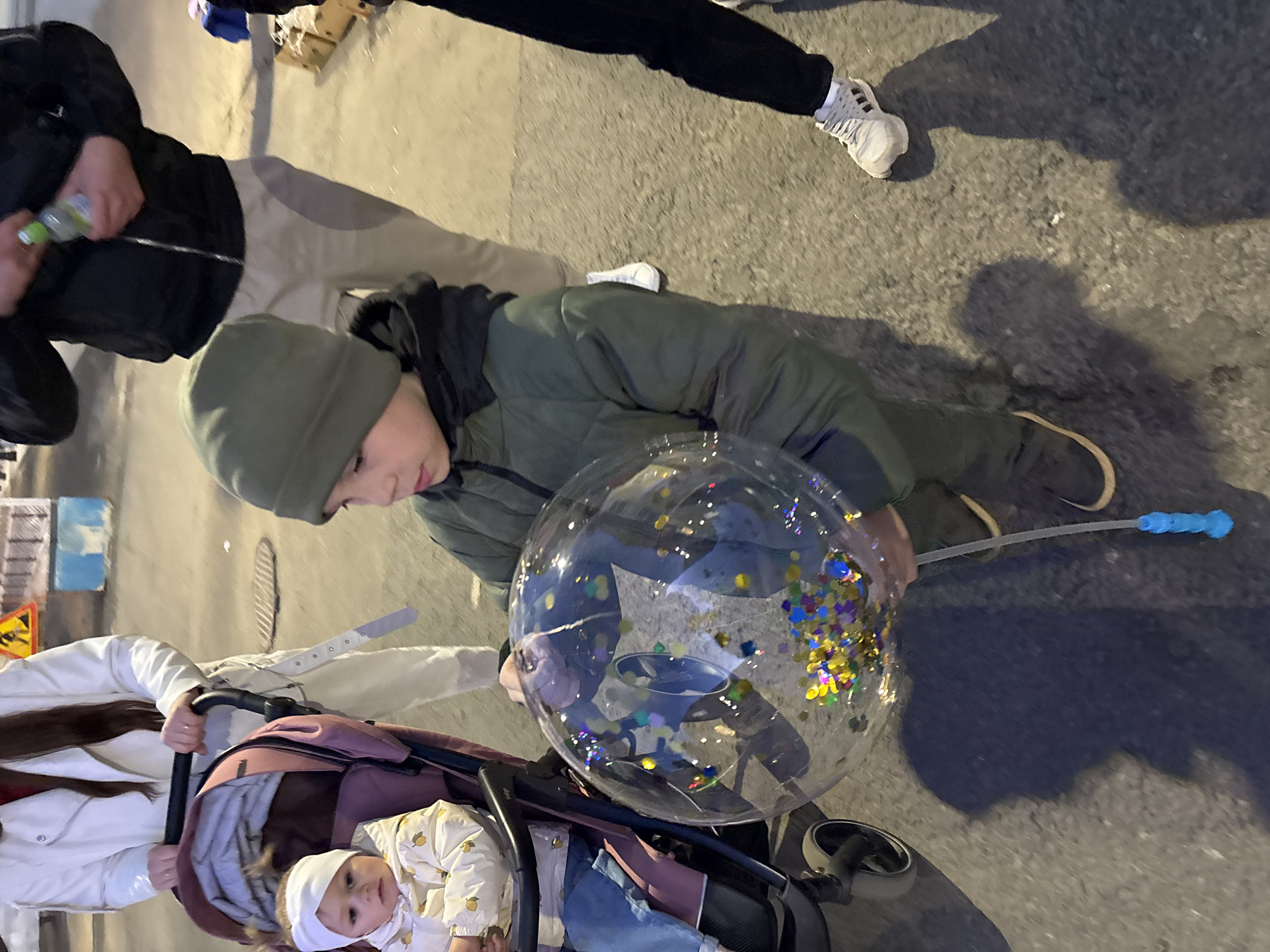 A sideways, high-angle shot shows a group of people on a city street in Rusanovo, Russia. An older child in a green jacket and hat is holding a transparent balloon filled with confetti, positioned above a baby in a stroller. A woman in a white jacket is pushing the stroller. Another person is visible in the background wearing a black jacket and light-colored pants, holding a water bottle. Also in the background, there's someone with black pants and white sneakers. The setting appears to be an urban sidewalk at night.