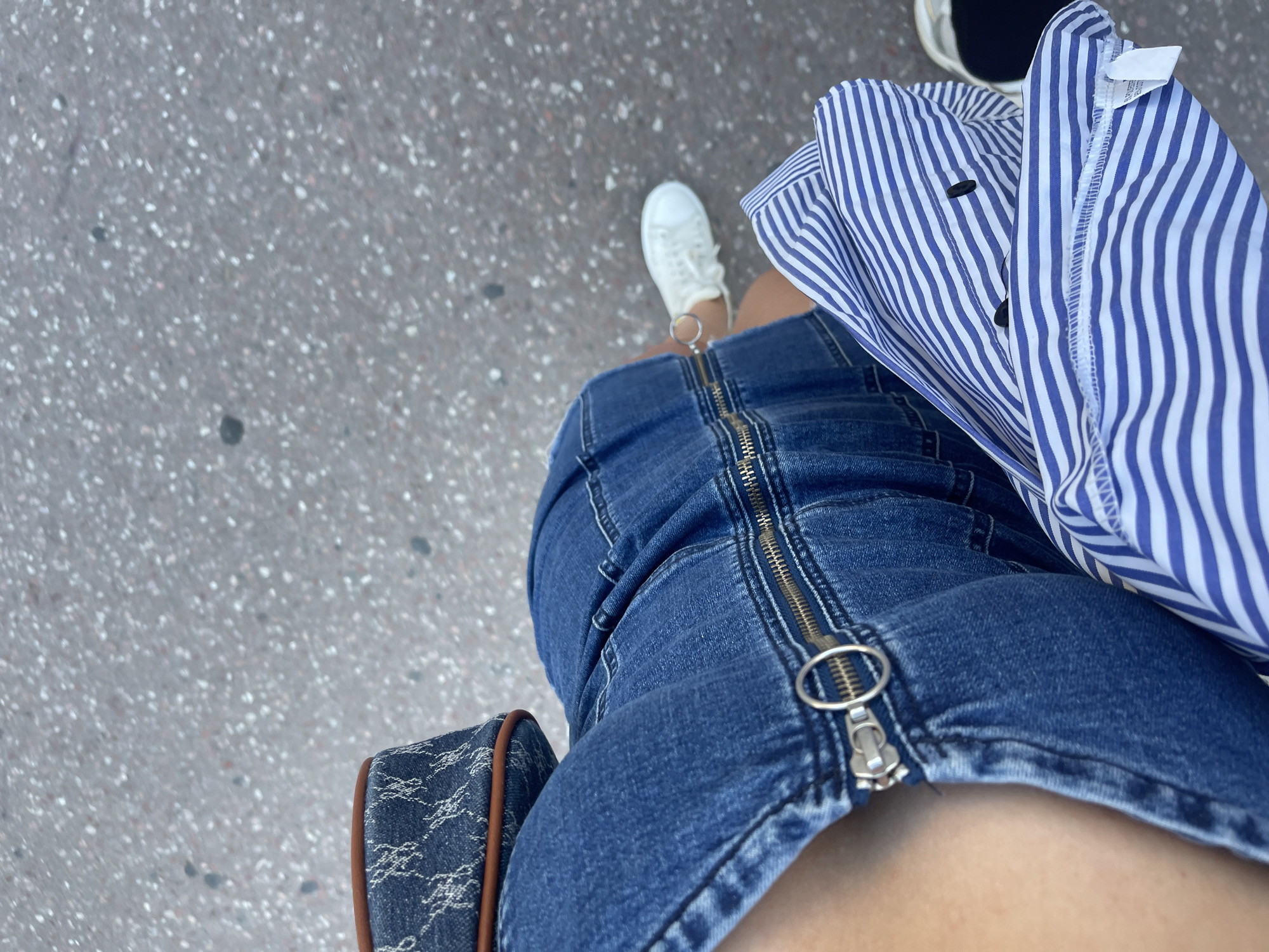 The image shows a person standing on a concrete surface, likely a sidewalk. The person is wearing a blue denim dress with a long, visible zipper down the front. They are also wearing a blue and white striped shirt and white sneakers. Part of a bag with a dark pattern and brown trim is visible near the bottom left of the frame.
The location cannot be determined to be in València, Spain from the image alone. More visual cues like distinctive architecture, street signs in Valencian or Spanish, or identifiable landmarks would be needed to confirm the location.