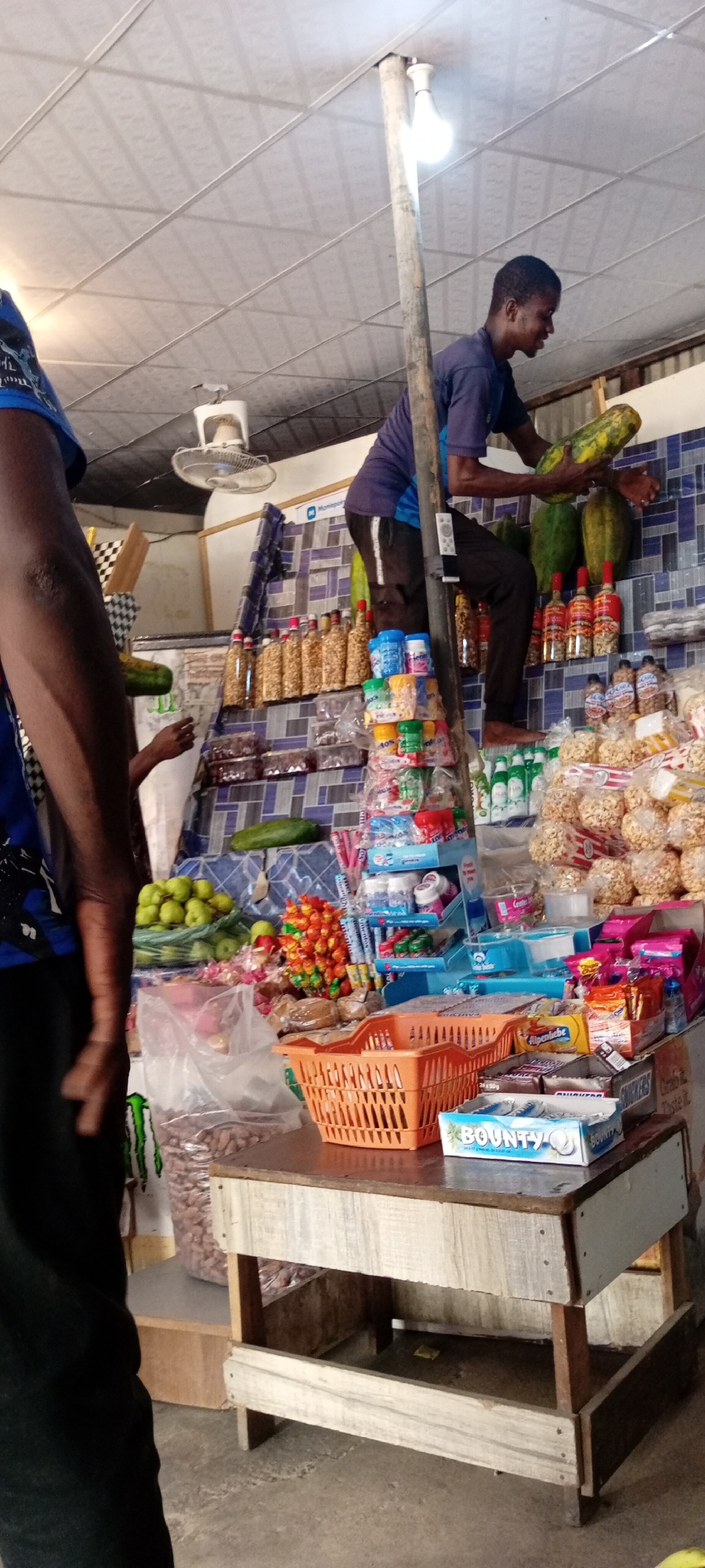 In Aviation Quarters, Nigeria, a young man is perched on a makeshift ladder made of wood, reaching for a large green papaya hanging from a shelf. He wears a dark purple t-shirt and dark pants. The shelves behind him are laden with various goods: bottles of what appear to be oil or sauce, bags of snacks, and assorted packaged food items, including Bounty chocolate bars. To the left, another person, only their arm and shoulder visible, is wearing a blue t-shirt. A white oscillating fan is mounted on the ceiling above. The overall setting is a small shop or market stall, with shelves constructed from wood and a tiled blue and white backsplash. The floor is concrete.