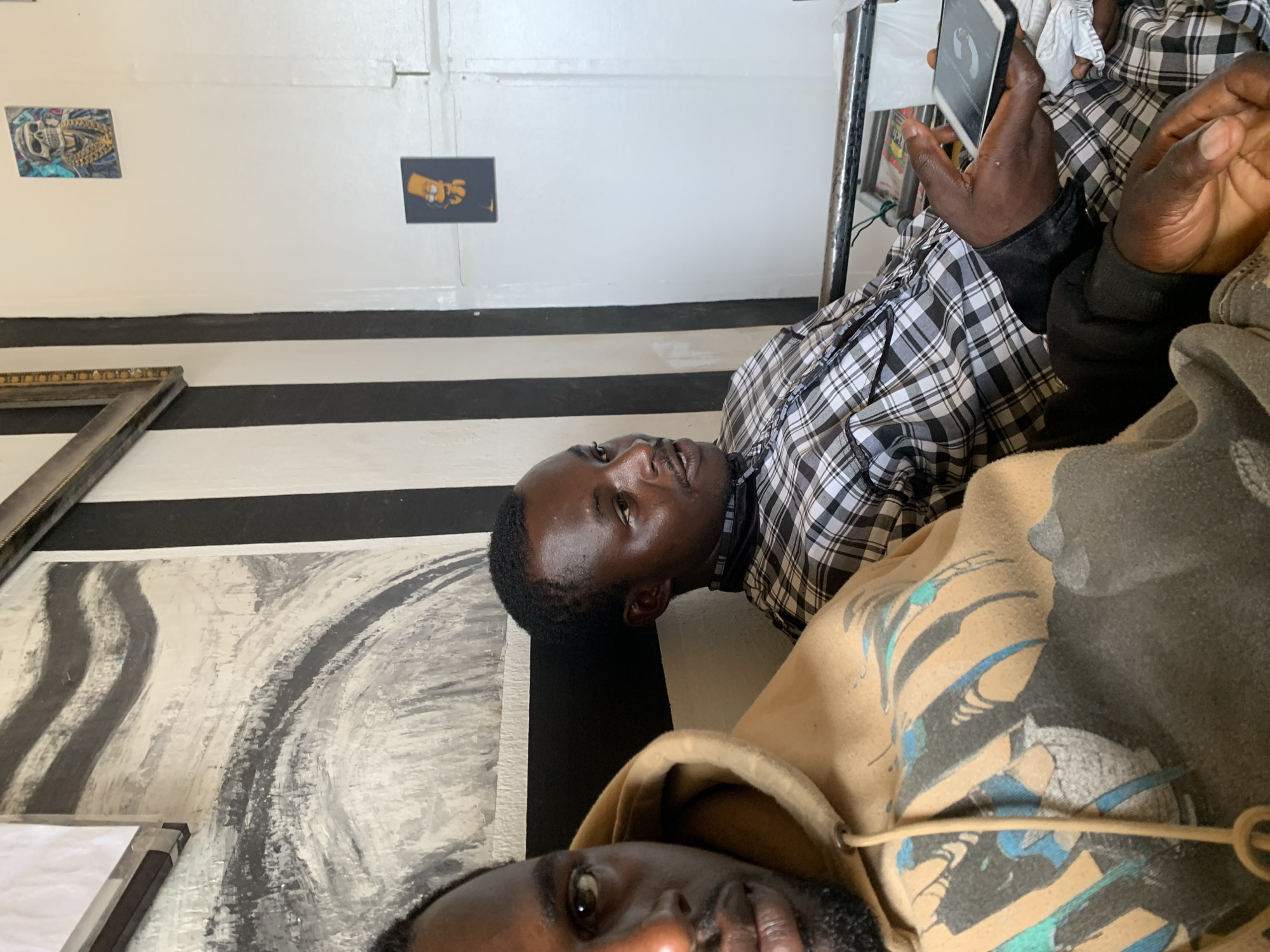 The image shows two dark-skinned men in an indoor setting. One man is wearing a tan hooded sweatshirt with a graphic design and the other is wearing a black and white checkered shirt. One of the men is holding a smartphone. Behind them is a wall painted with black and white horizontal stripes. Above the striped wall, there are two small posters and a framed painting, which appears to be an abstract design. The location is likely in Jos, Nigeria, based on cultural context and assumptions.