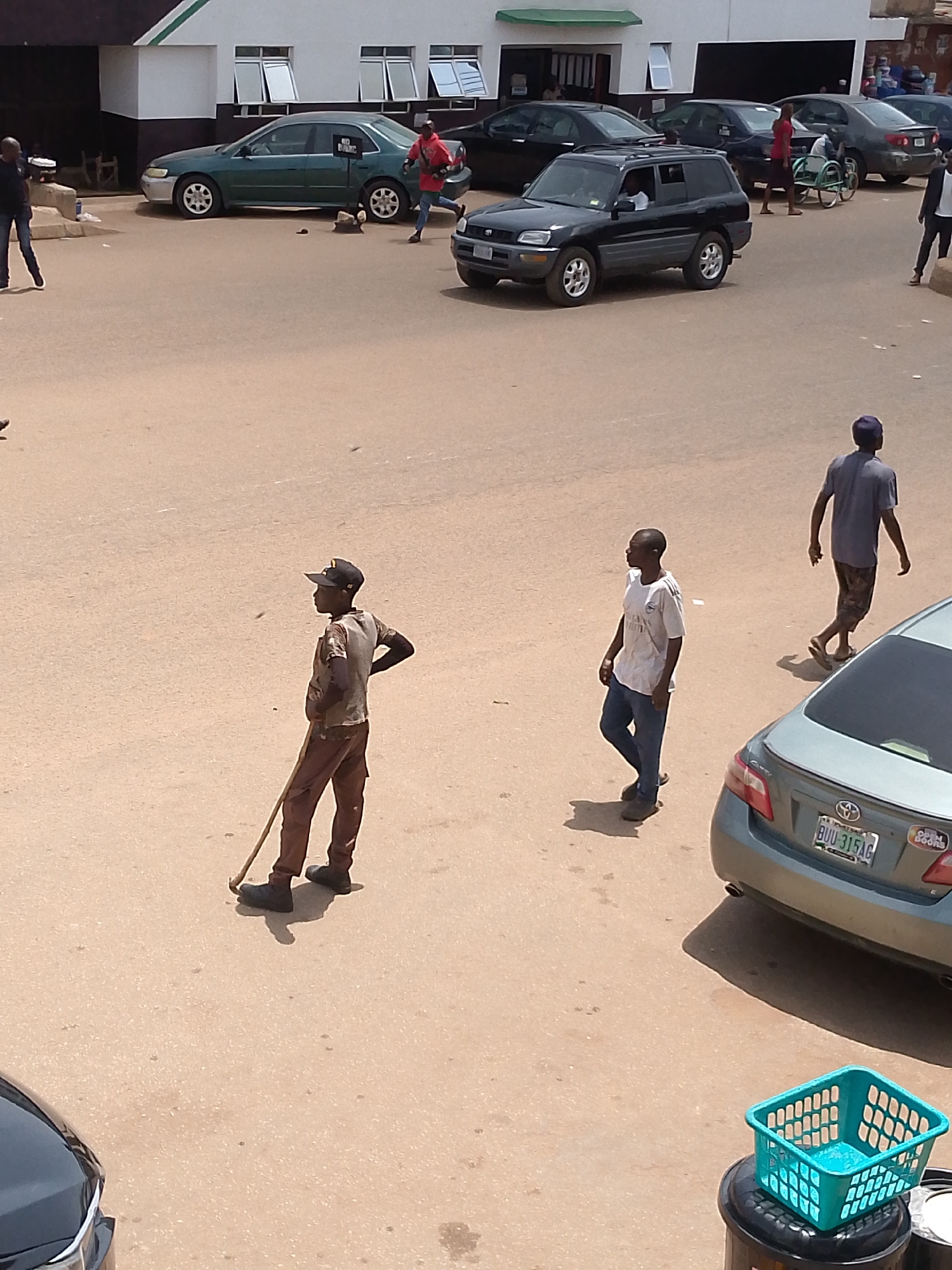 This media file captures a street scene in Bukuru, Nigeria, under what appears to be bright daylight, suggesting a warm and sunny day. The setting is an urban environment with a building in the background featuring windows and a green trim. The ground is paved with a light-colored, sandy material, typical of many Nigerian streets.
Several people are present in the scene. In the foreground, a young person wearing a cap, a light-colored patterned shirt, and brown trousers stands with one hand on their hip, holding a long, thin object that resembles a broom or a stick. Further back, another person in a white t-shirt and blue jeans walks, looking to the left. In the distance, a person in a blue t-shirt and patterned shorts walks away from the camera.
Various vehicles are visible, including a dark SUV, a green sedan, and a lighter colored sedan in the foreground with a visible license plate reading "BUU 315AG". A blue plastic basket sits atop a black trash can in the lower right corner of the image.
The overall impression is of an ordinary day in a town, with people going about their activities. There are no overtly dramatic events unfolding. The lighting suggests it's daytime, likely mid-morning to afternoon, due to the strong shadows. The environment appears open and dusty.