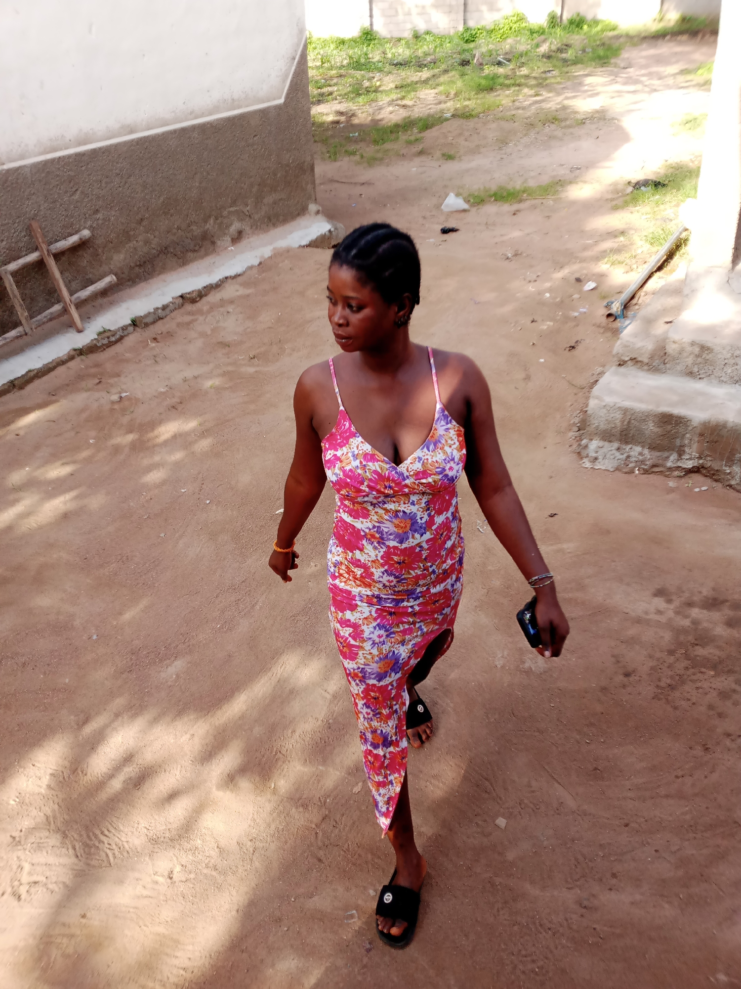 In Jauro Yinu, Nigeria, a Black woman with braided hair walks outdoors on a dirt path. She wears a pink floral-patterned dress with spaghetti straps and black slide sandals. Her right hand holds a dark-colored mobile phone, and she has a bracelet on her left wrist. The environment appears to be a residential area with a textured wall of a building to the left and a concrete structure with stairs to the right. The ground is predominantly sandy and dusty, with some scattered debris. Sunlight casts shadows on the ground, indicating it is daytime.